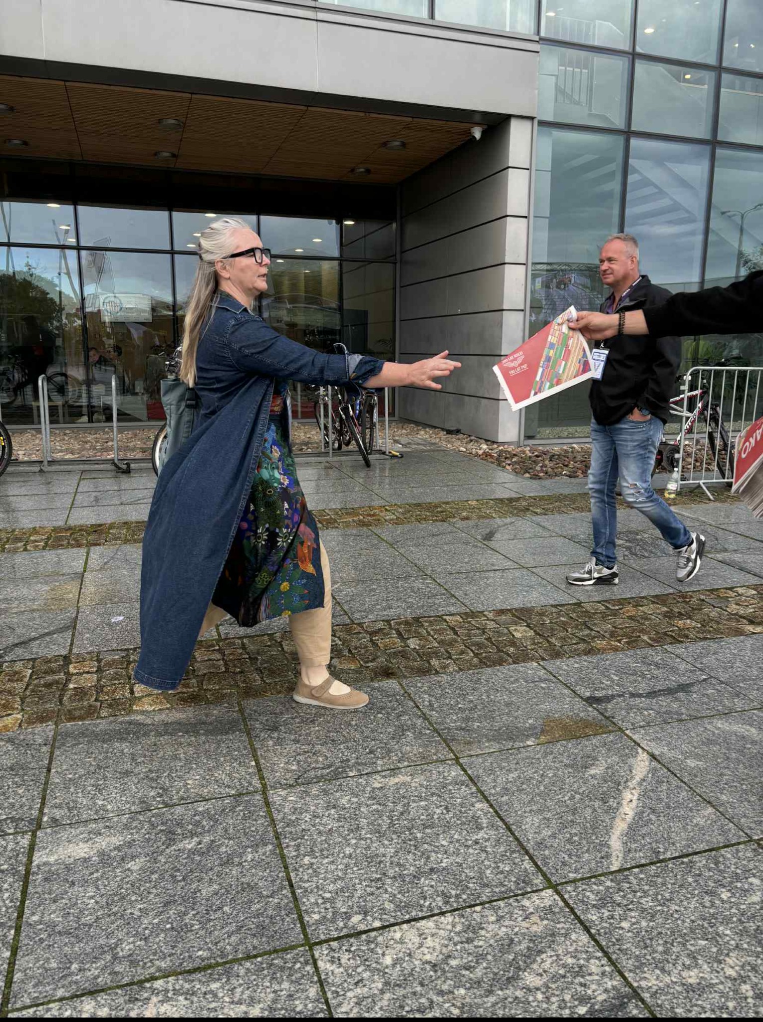 Kobieta w dżinsowej sukience wyciąga ręce po ulotkę wręczaną przez mężczyznę na tle nowoczesnego budynku z przeszkloną fasadą. Wydarzenie plenerowe.