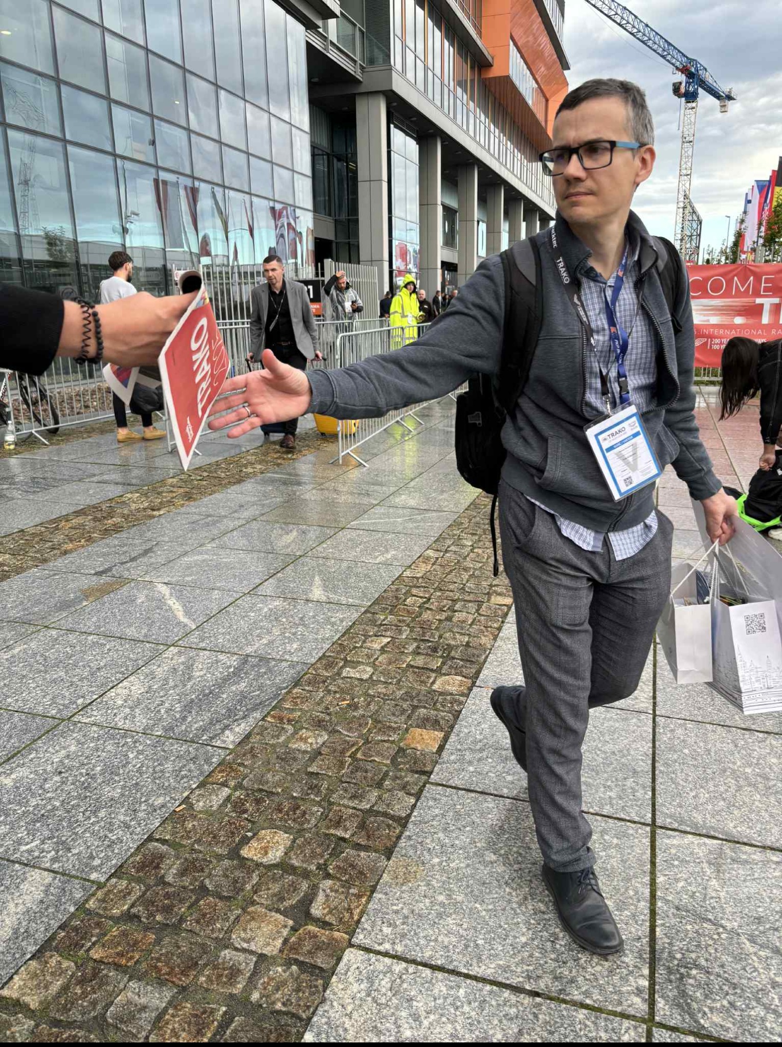 Mężczyzna w okularach i szarej bluzie odbiera ulotkę na tle nowoczesnego budynku z napisem 'WELCOME'. Widoczny żuraw budowlany w tle.