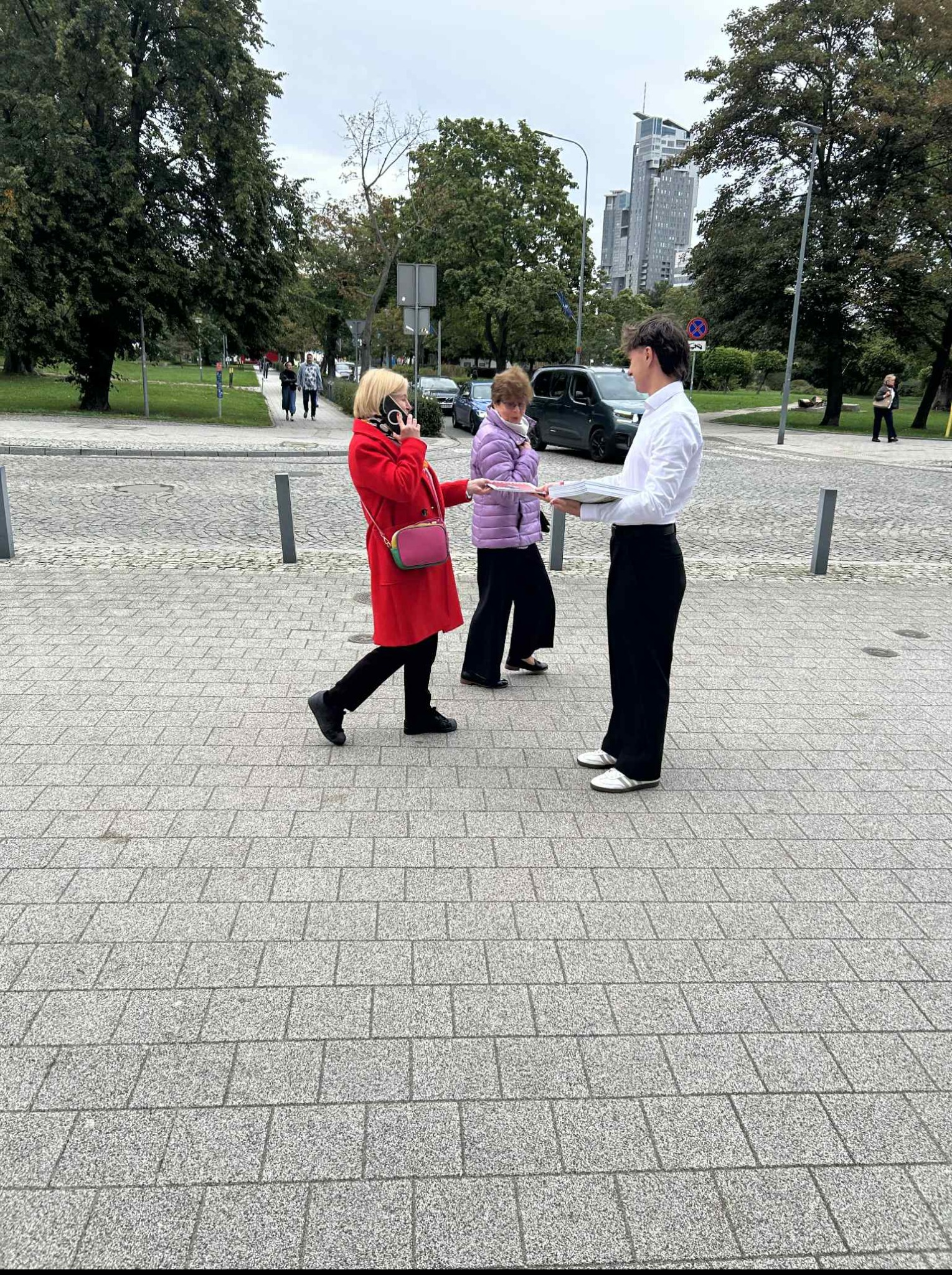 Promotor w białej koszuli wręcza ulotki kobiecie w czerwonym płaszczu, która rozmawia przez telefon. W tle budynki i drzewa.