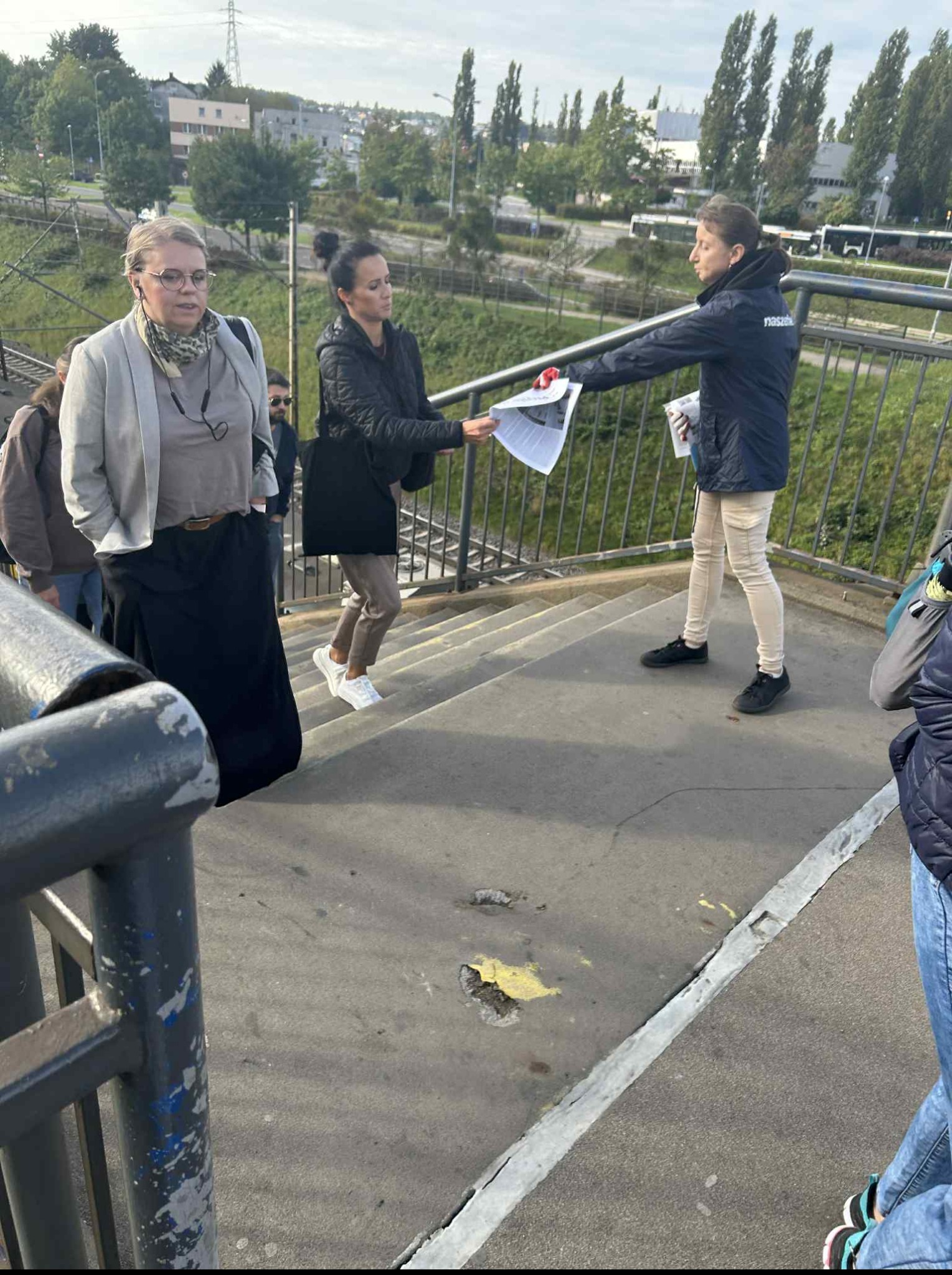 Rozdawanie ulotek na schodach wiaduktu. Osoba w kurtce z napisem 'nasze' wręcza ulotki przechodniom. Widoczne budynki w tle i fragmenty innych osób.
