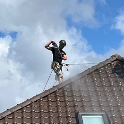 Dbamy o Dach - Pracownik w kasku, z zabezpieczeniem linowym, czyści dach z brązowej dachówki za pomocą myjki ciśnieniowej. Widoczne okno dachowe i błękitne niebo z chmurami.