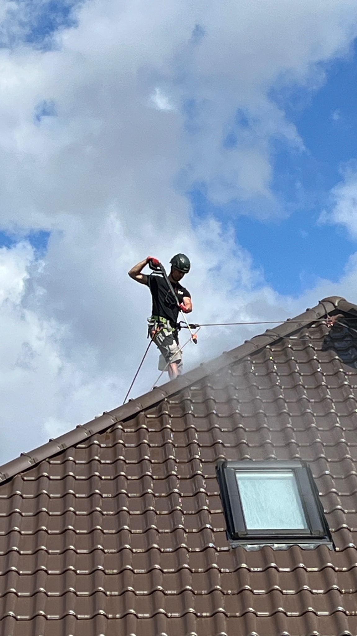 Pracownik w kasku, z zabezpieczeniem linowym, czyści dach z brązowej dachówki za pomocą myjki ciśnieniowej. Widoczne okno dachowe i błękitne niebo z chmurami.