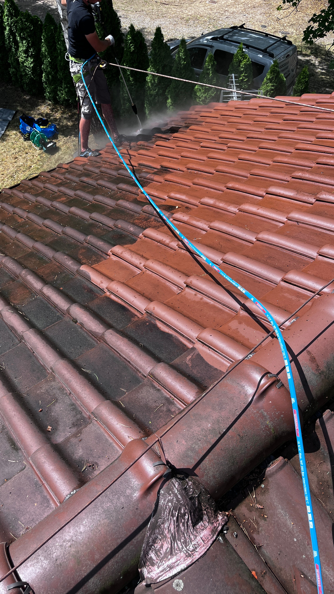 Częściowo umyty dach z czerwonej dachówki, widoczny pracownik w uprzęży i lanca ciśnieniowa. Połowa dachu ciemna, zabrudzona, druga połowa czysta, umyta.