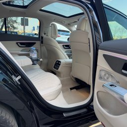 MAT VIP TRANSPORT - Luksusowe wnętrze samochodu z jasną skórzaną tapicerką, otwartymi drzwiami i panoramicznym dachem. Widoczne butelki wody Evian na podłokietniku.