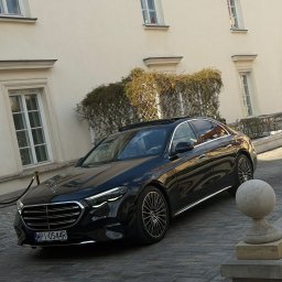 MAT VIP TRANSPORT - Elegancki, czarny Mercedes-Benz S-klasa z warszawskimi tablicami rejestracyjnymi zaparkowany na brukowanej ulicy przed budynkiem z jasną elewacją i ozdobną roślinnością.