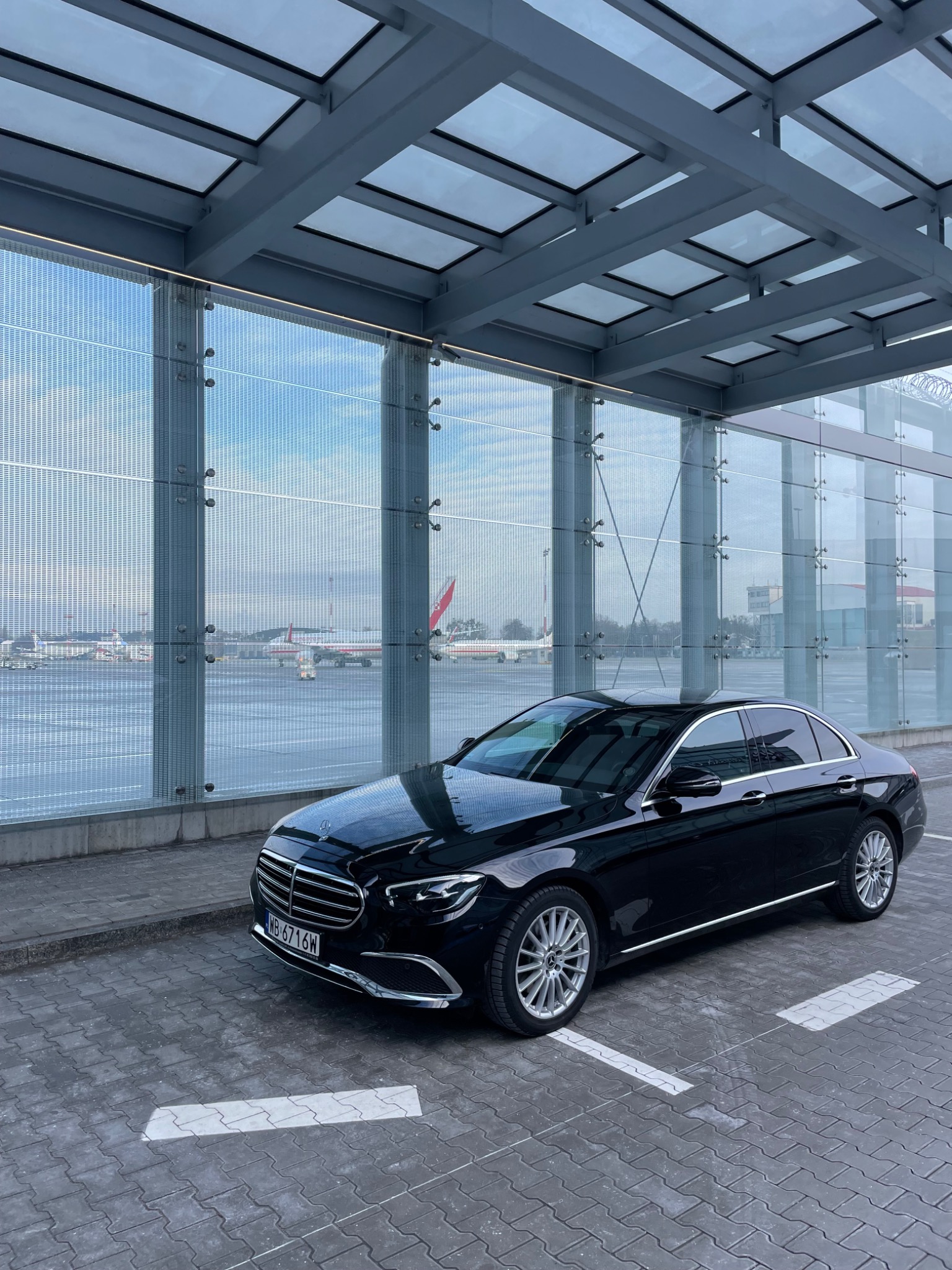 Czarna limuzyna Mercedes klasy E na parkingu lotniska, z widokiem na samoloty za szklaną fasadą terminalu. Elegancki pojazd gotowy do transportu pasażerów.