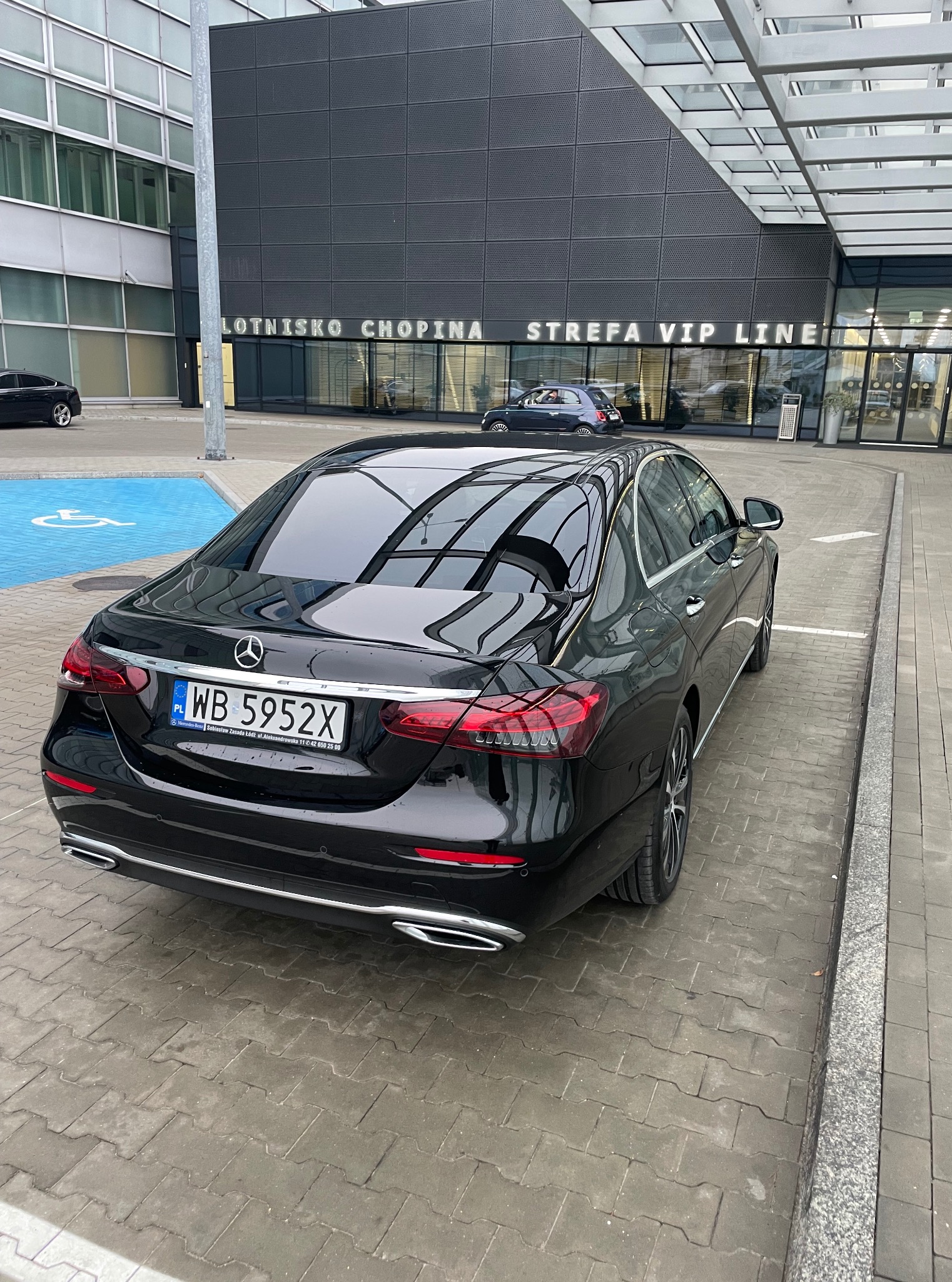 Czarny Mercedes-Benz E-Klasa z polską rejestracją WB 5952X na tle Lotniska Chopina, Strefa VIP Line. Widok z tyłu, lekki kąt. Reprezentacyjny charakter.