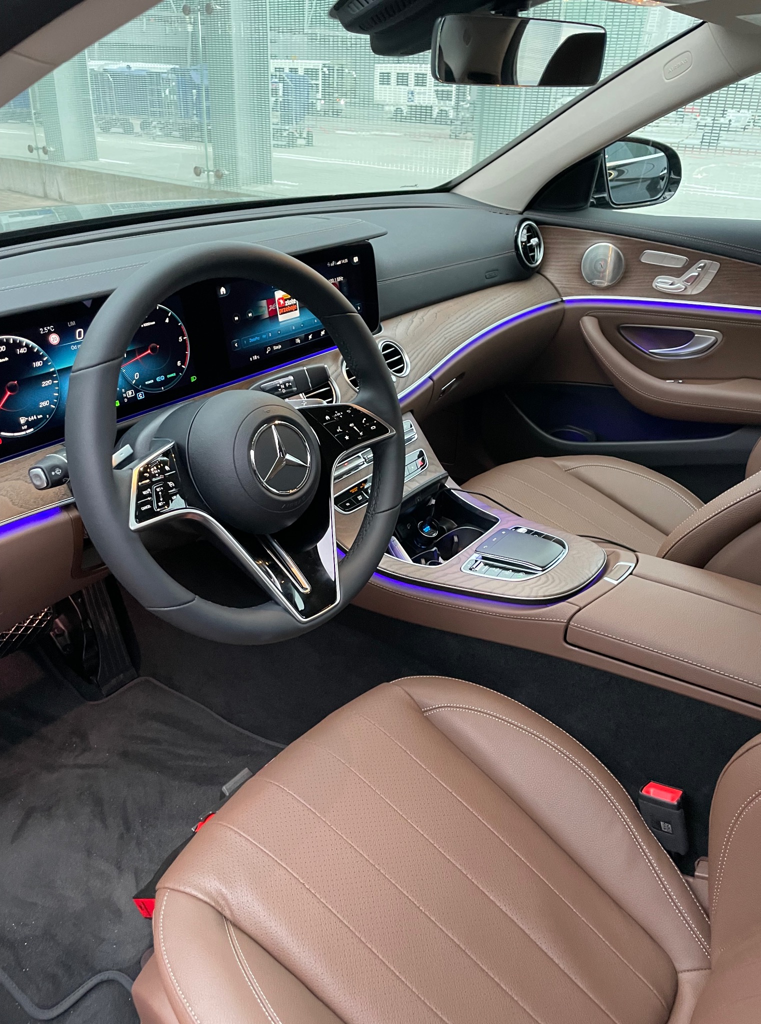 Luksusowe wnętrze samochodu Mercedes z brązową skórzaną tapicerką, drewnianymi akcentami i subtelnym, fioletowym podświetleniem ambientowym. Widoczna kierownica z logo.