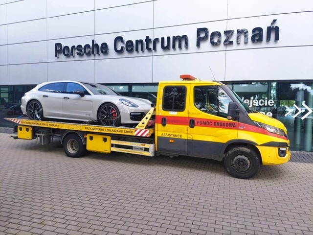 Żółta laweta z Porsche Panamera GTS na tle Porsche Centrum Poznań. Pomoc drogowa, auto na platformie, widoczne napisy informacyjne na pojeździe.