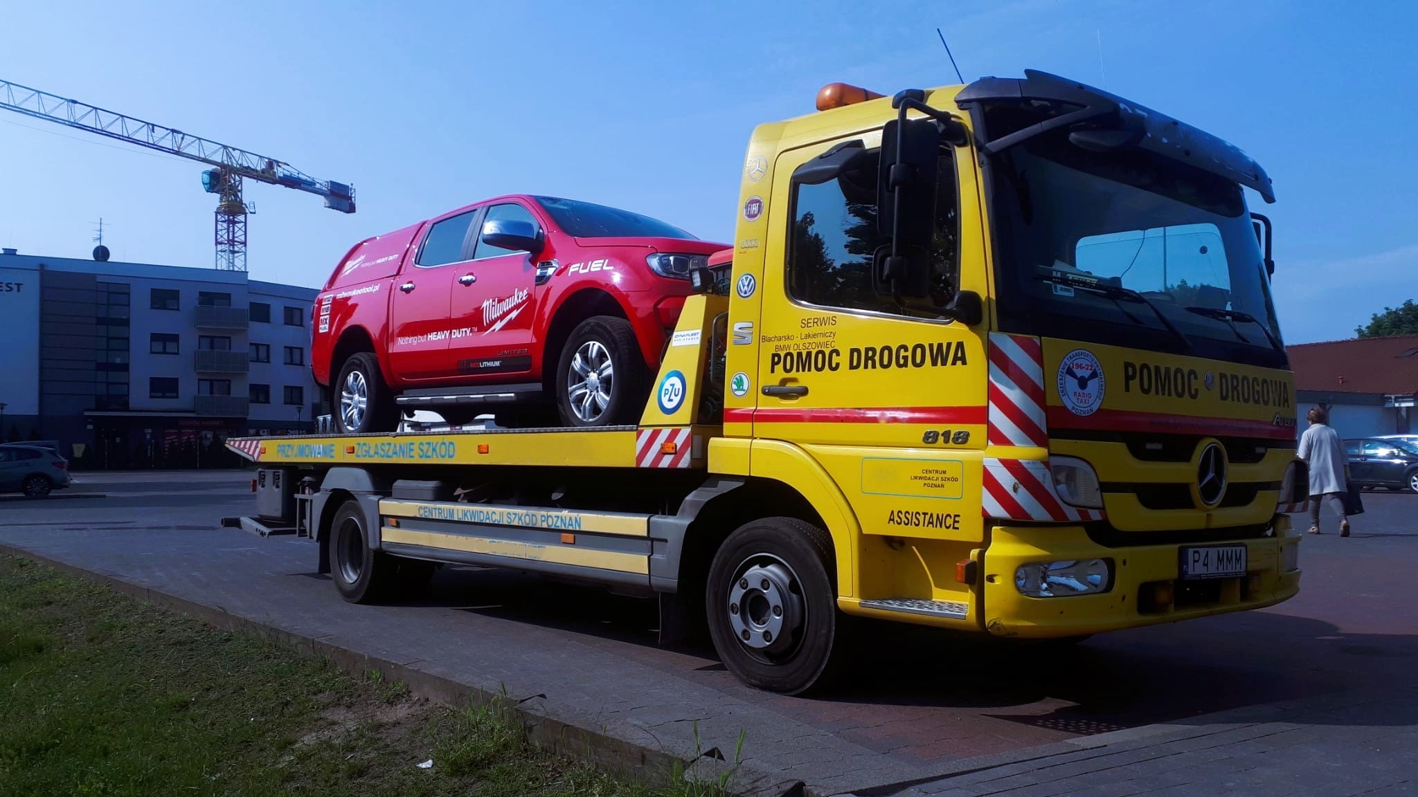 Żółta laweta Mercedes z czerwonym pickupem Milwaukee na platformie, na tle budynku i dźwigu. Widoczne logo firm na samochodzie.