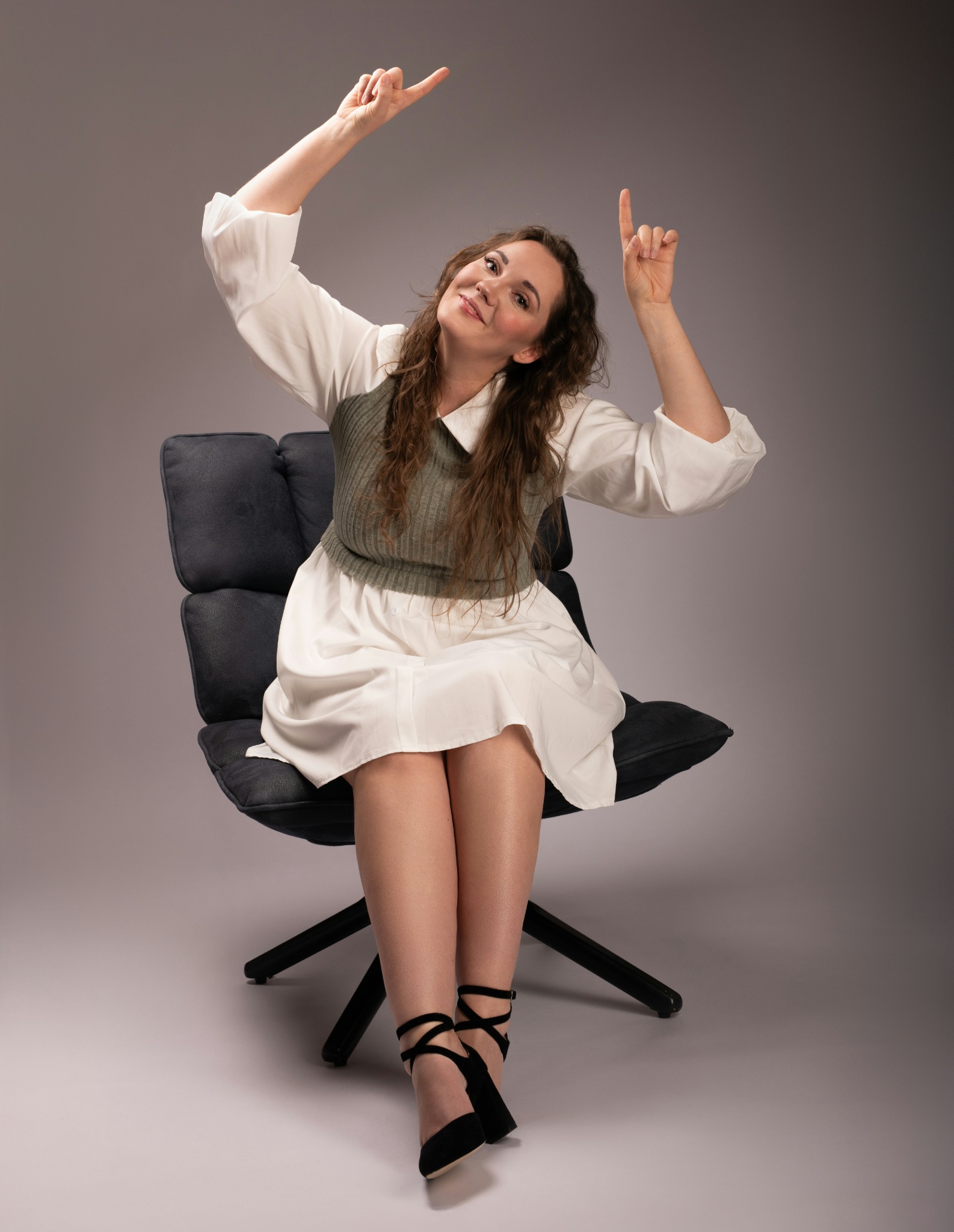 Uśmiechnięta kobieta siedzi na krześle, wskazując palcami do góry. Sesja w studio na szarym tle. Czarno-biała sukienka i czarne buty na obcasie.