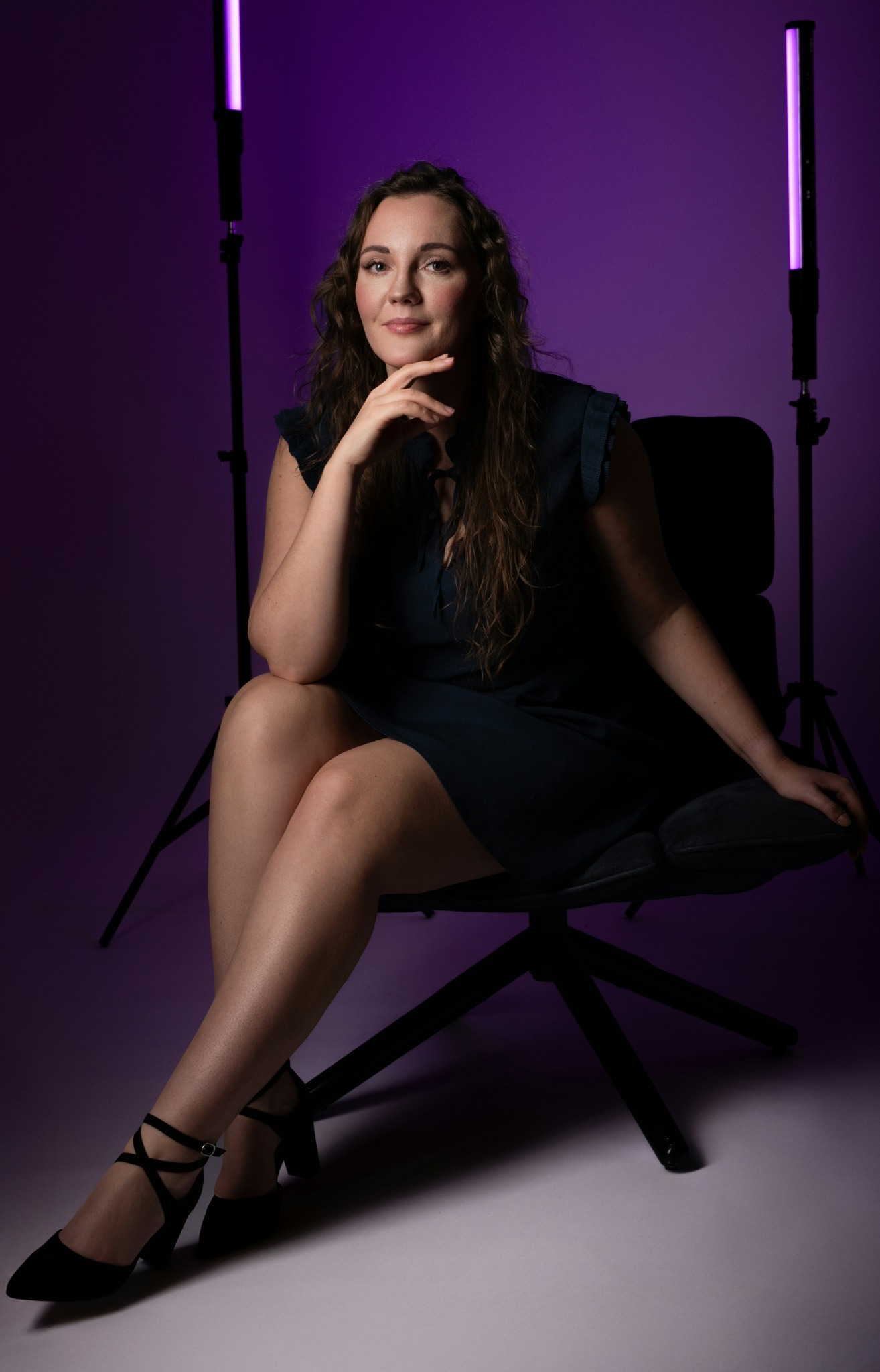 Portret kobiety siedzącej na krześle na fioletowym tle, oświetlonej lampami LED. Kobieta ma długie włosy i jest ubrana w sukienkę. Uwagę przyciągają czarne buty na obcasie.
