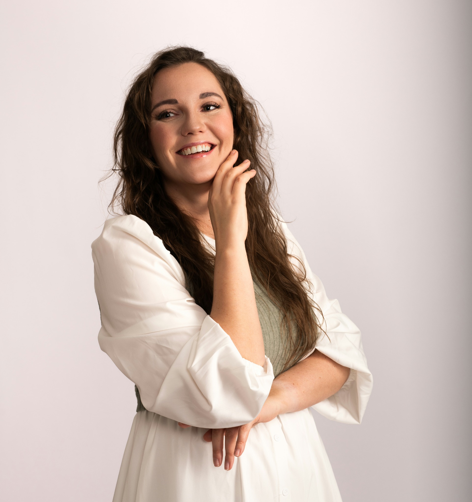 Uśmiechnięta brunetka w białej sukience pozuje z dłonią przy twarzy na jasnym tle. Portret wizerunkowy z subtelnym oświetleniem i naturalnym makijażem.