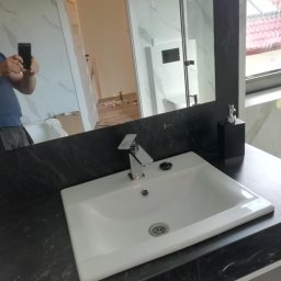 Andrzej Ludwig - Biała umywalka nablatowa na czarnym blacie w wyremontowanej łazience w Słupsku. W lustrze odbicie części pomieszczenia i osoby robiącej zdjęcie.