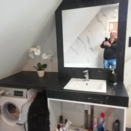 Andrzej Ludwig - Łazienka na poddaszu z umywalką, blatem z czarnego kamienia i pralką. W lustrze odbija się osoba robiąca zdjęcie. Uwagę zwraca nietypowy kształt lustra.