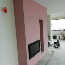 Andrzej Ludwig - Wnęka kominkowa w zabudowie z płyt kartonowo-gipsowych, z czarnym wkładem kominkowym. Widoczne narzędzia i materiały budowlane w tle, surowy stan wykończenia wnętrza.