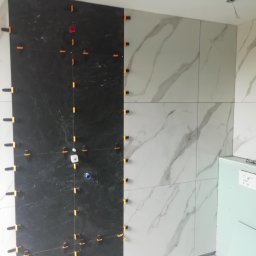 Andrzej Ludwig - Układanie płytek: czarne i białe marmurowe płytki na ścianie, z pomarańczowymi klipsami poziomującymi, widoczne fragmenty podłogi i narzędzi glazurniczych.