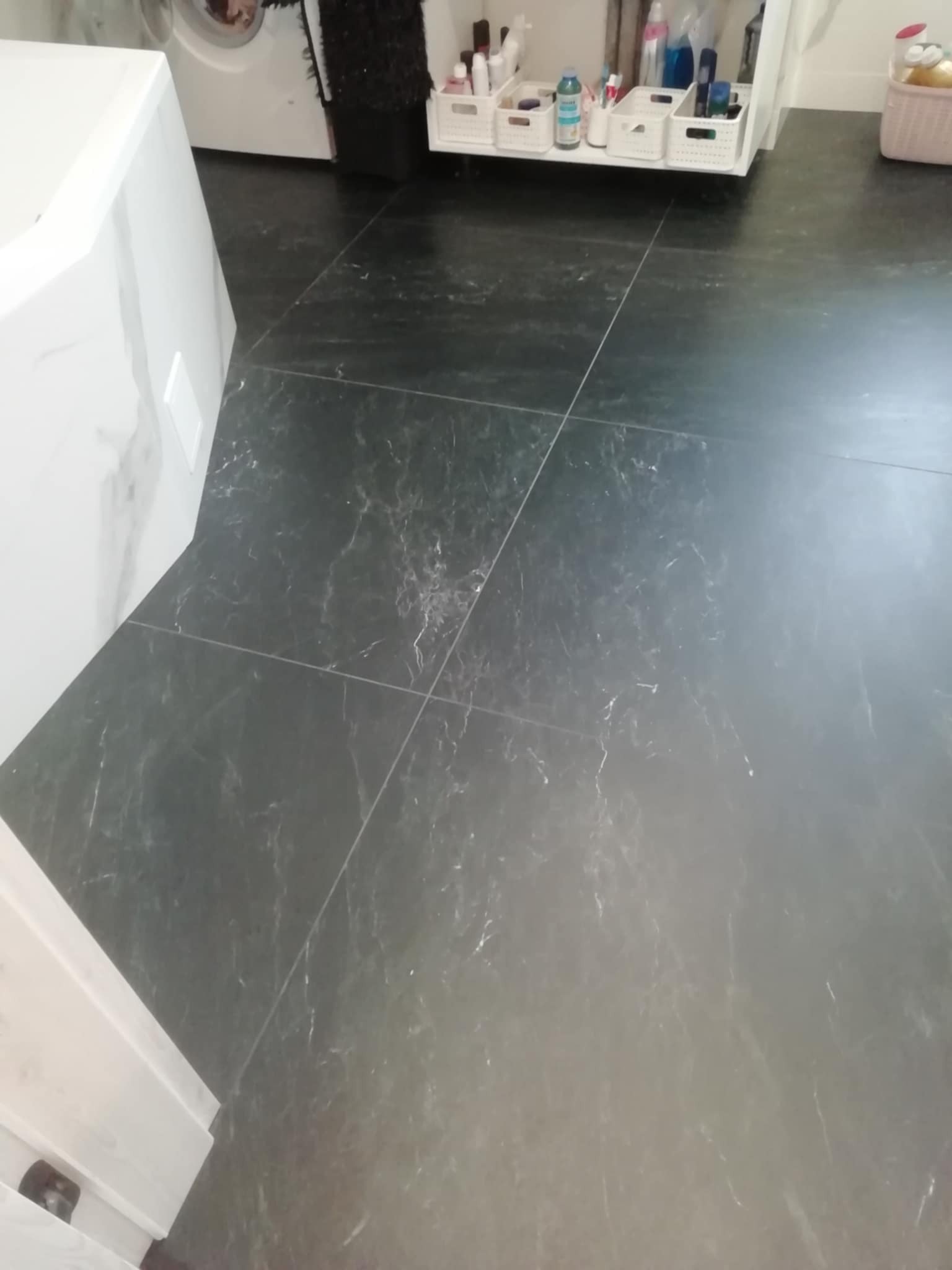 Ciemna, marmurkowa podłoga z płytek w łazience, widok z góry, fragment pralki i szafki z kosmetykami w tle. Minimalistyczny design, czyste fugi, nowoczesne wykończenie.