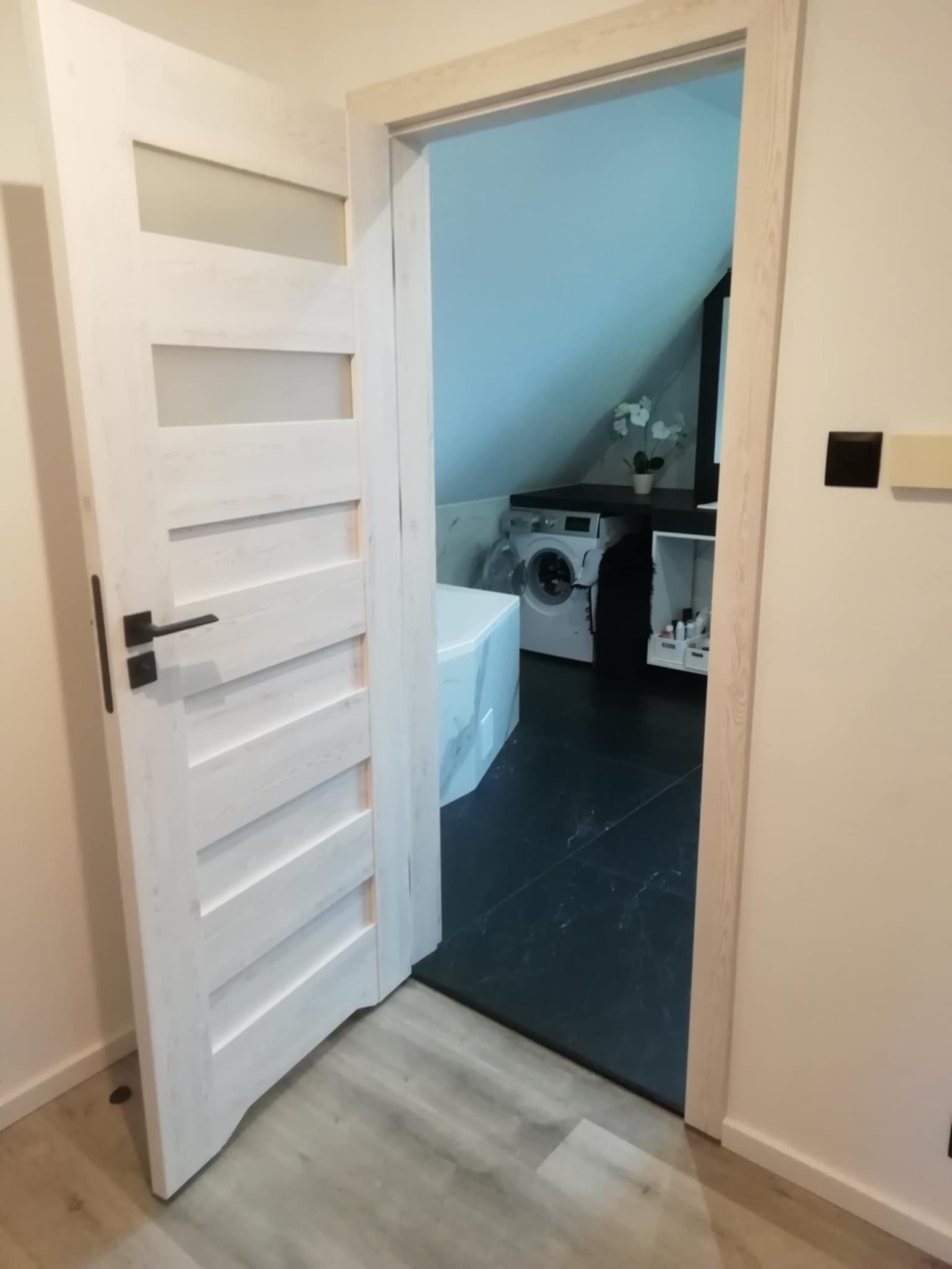 Otwarta, biała, drewniana drzwi z przeszkleniami, czarna klamka. W tle łazienka na poddaszu z pralką, blatem i marmurową obudową wanny.