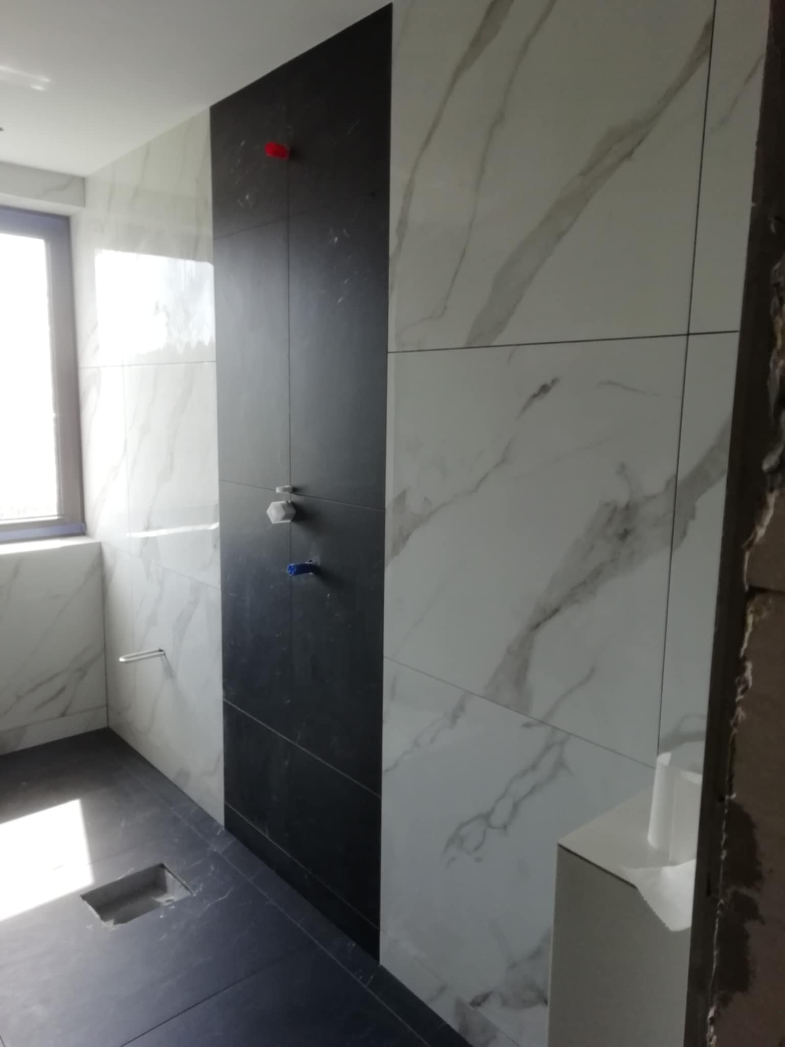 Nowoczesna łazienka z czarną ścianą prysznicową i marmurowymi płytkami. Widoczne baterie i odpływ liniowy. Minimalistyczny design z dbałością o detale.