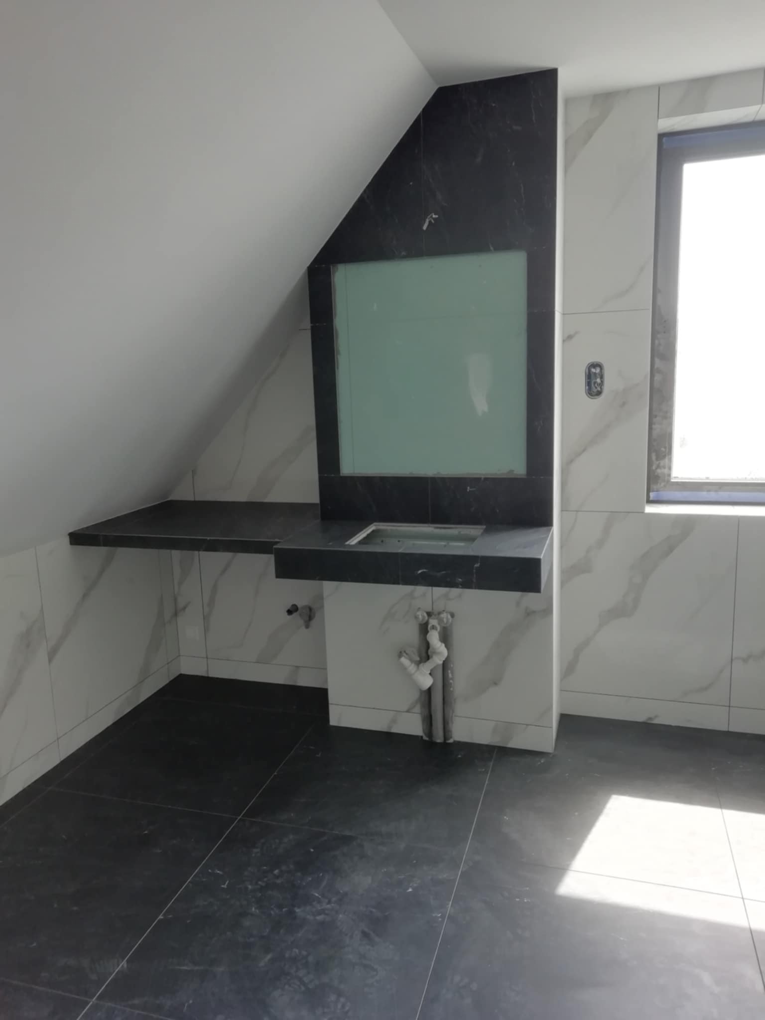 Łazienka na poddaszu z umywalką i lustrem w czarnej oprawie. Białe płytki imitujące marmur na ścianach, czarna podłoga. Widoczne rury odpływowe.