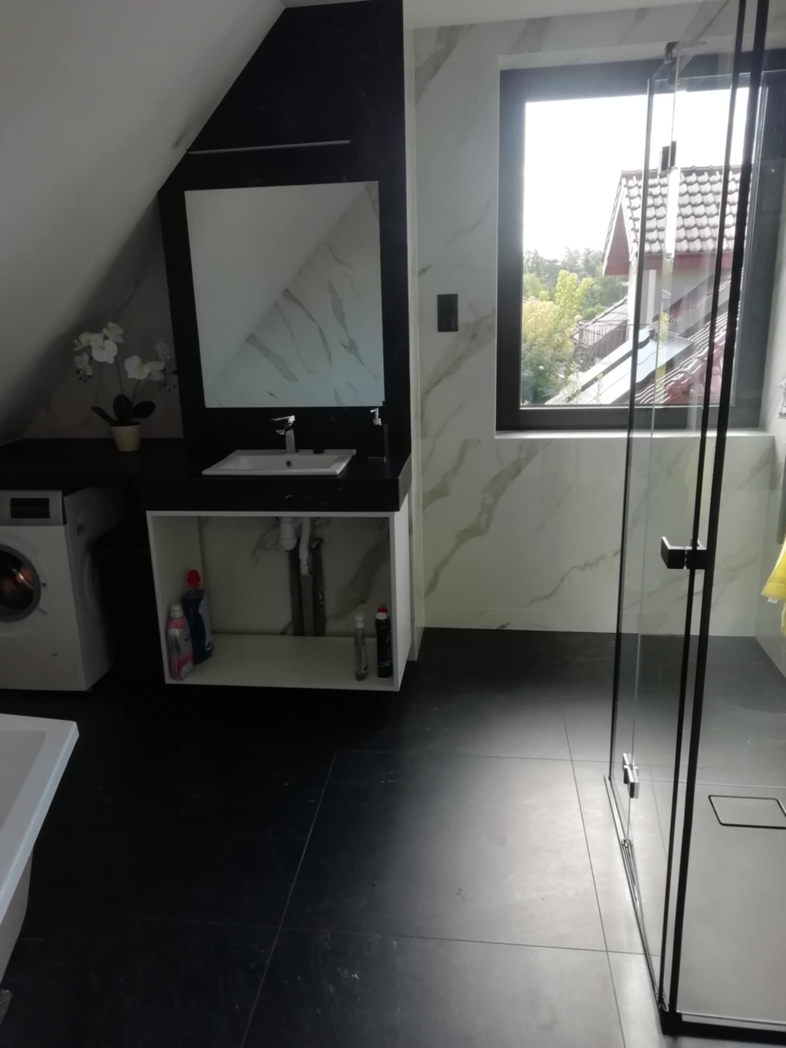 Nowoczesna łazienka na poddaszu: czarne płytki, umywalka z szafką, kabina prysznicowa z czarnymi ramami i widok z okna na dom z czerwoną dachówką.