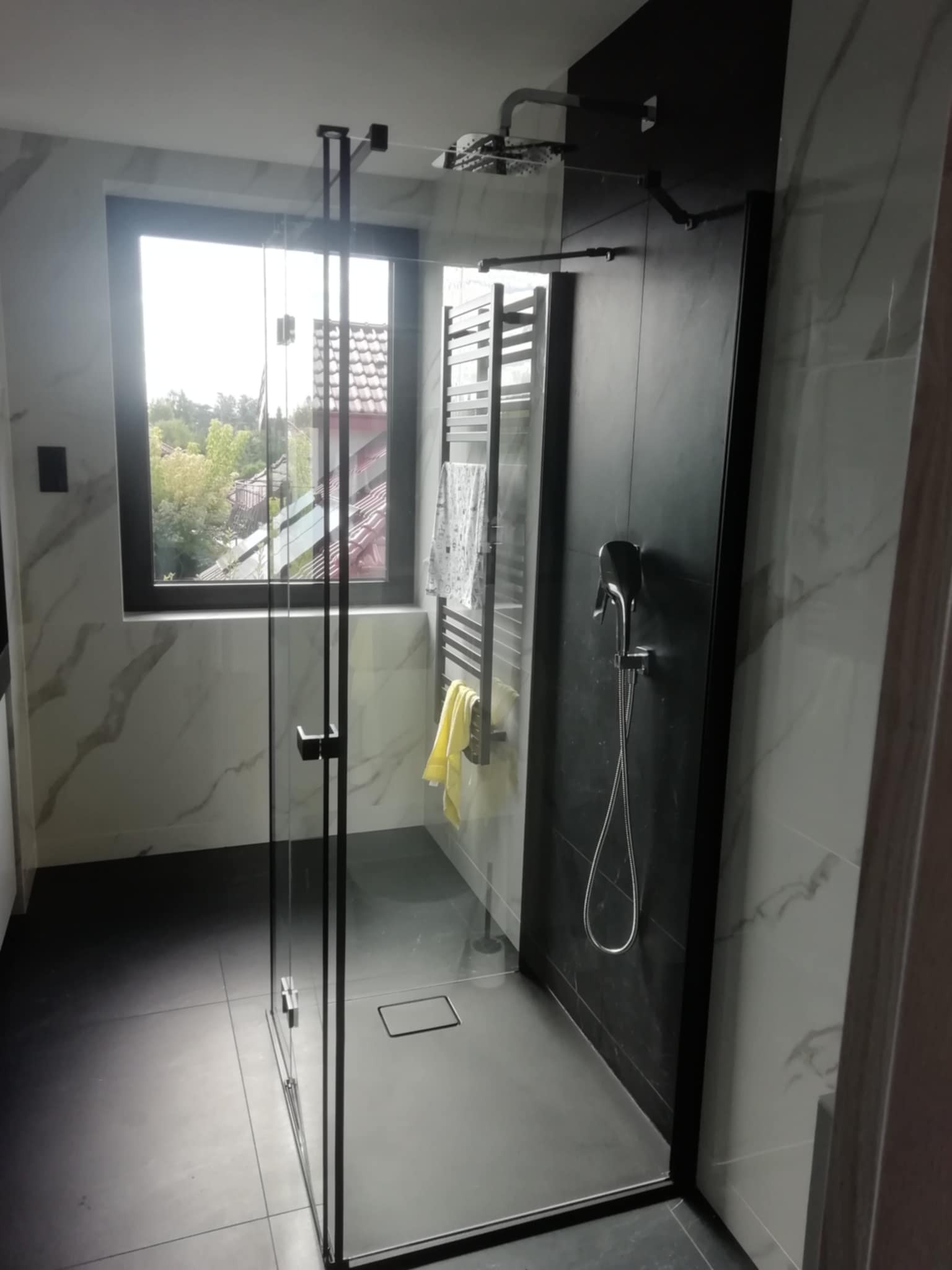 Nowoczesna łazienka z kabiną typu walk-in, czarne akcenty, marmurowe płytki, okno z widokiem na zieleń. Minimalistyczny design z grzejnikiem drabinkowym i deszczownicą.