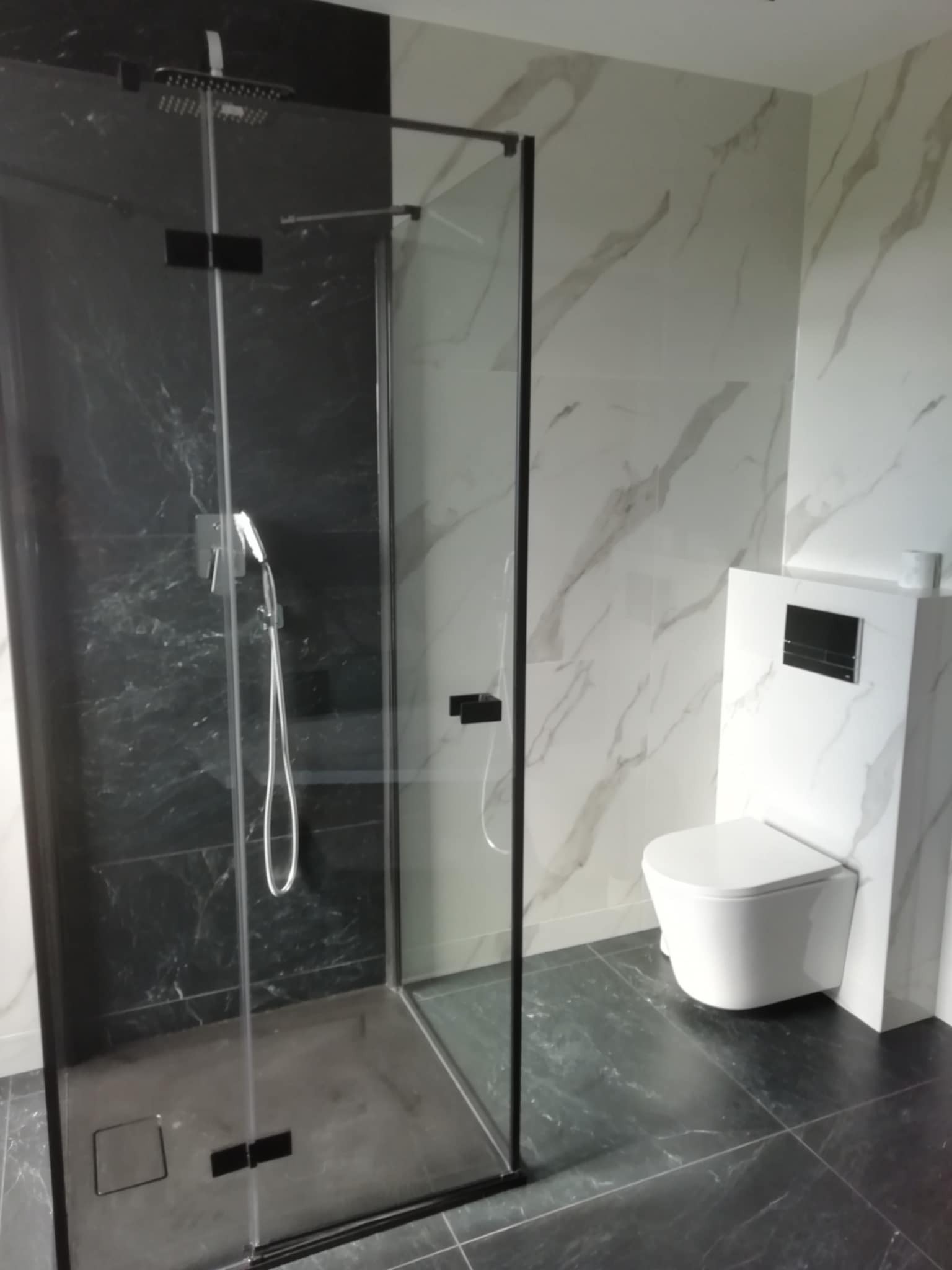 Nowoczesna łazienka z kabiną walk-in i białą ceramiką. Czarne płytki imitujące marmur na podłodze i ścianie kabiny, białe marmurowe płytki na pozostałej części ścian.