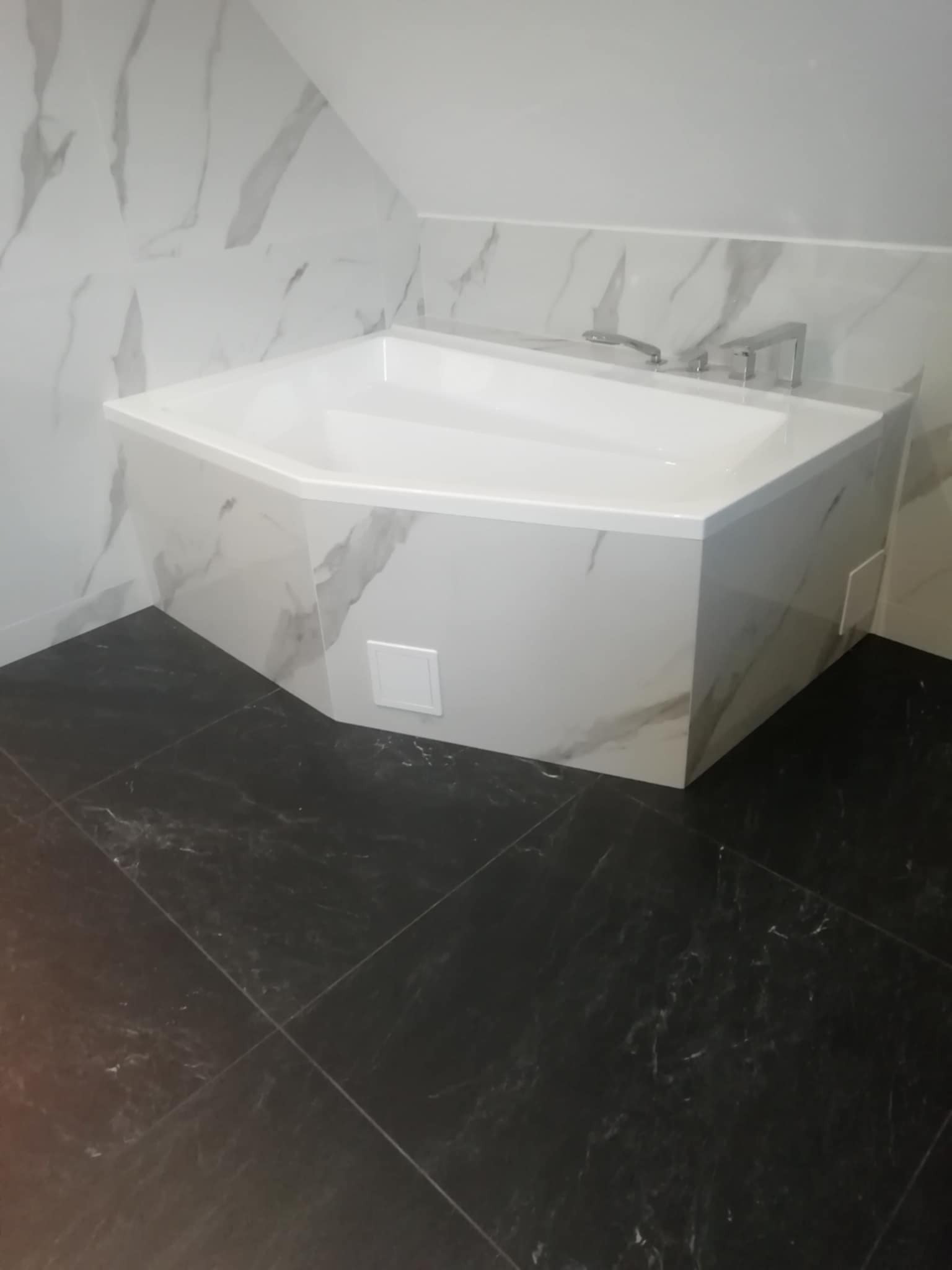 Biała narożna wanna obudowana płytkami imitującymi marmur, z czarną marmurową podłogą w wykończonej łazience na poddaszu.