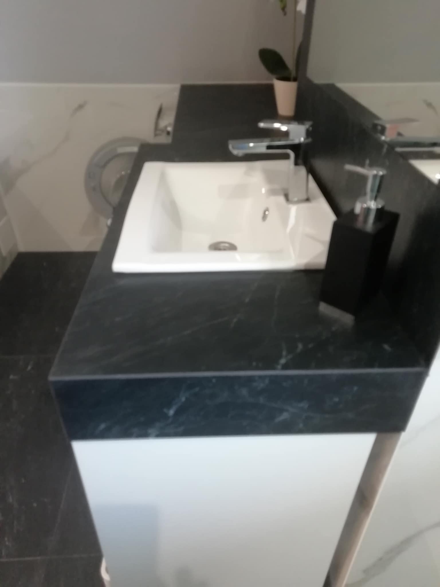 Biała umywalka z chromowaną baterią na blacie z czarnego marmuru w łazience, obok czarny dozownik na mydło. Widoczny fragment szafki pod umywalką.