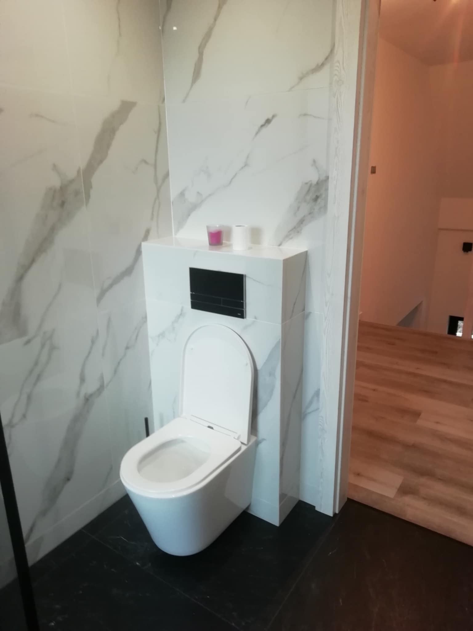 Nowoczesna łazienka po remoncie: podwieszana toaleta z czarnym panelem na tle białych płytek imitujących marmur i czarnej podłogi. Widok na otwarte drzwi.