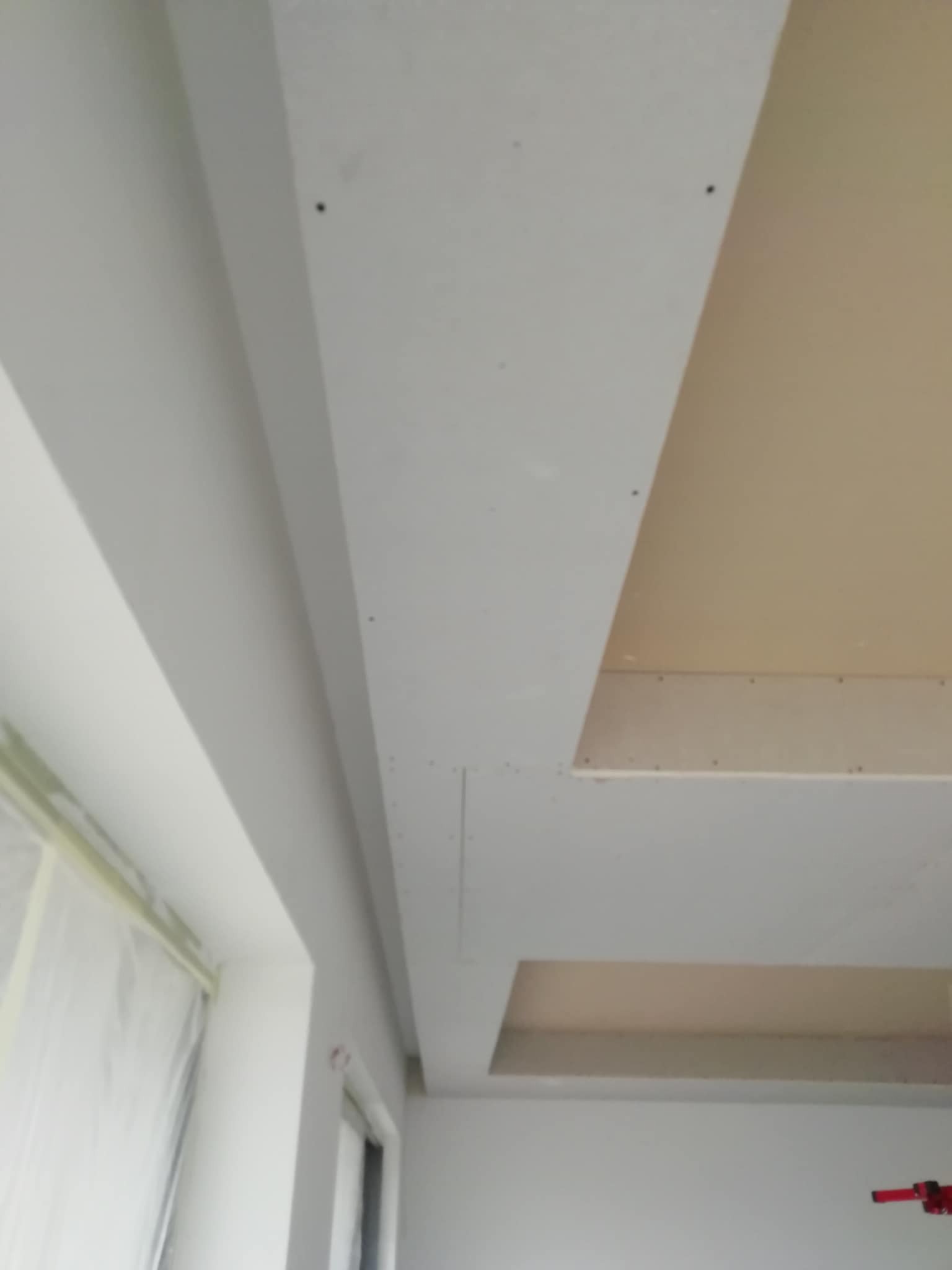 Sufit podwieszany z płyt gipsowo-kartonowych, częściowo wykończony, widoczne łączenia płyt i przygotowane do szpachlowania, okno zabezpieczone folią malarską.