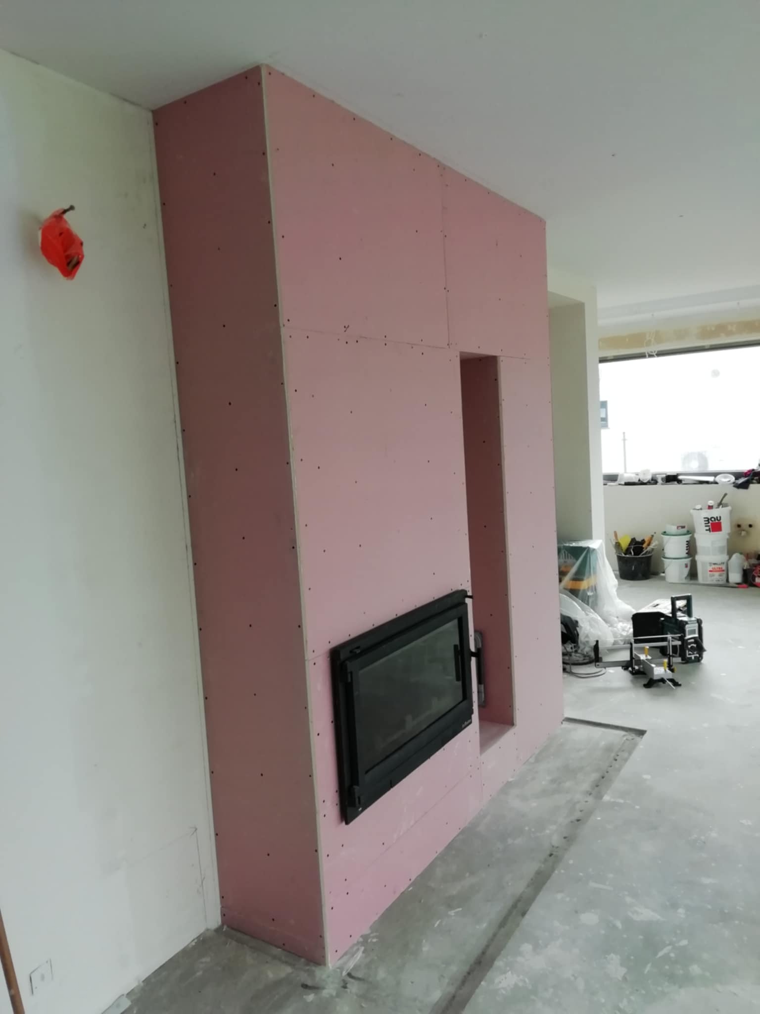 Wnęka kominkowa w zabudowie z płyt kartonowo-gipsowych, z czarnym wkładem kominkowym. Widoczne narzędzia i materiały budowlane w tle, surowy stan wykończenia wnętrza.