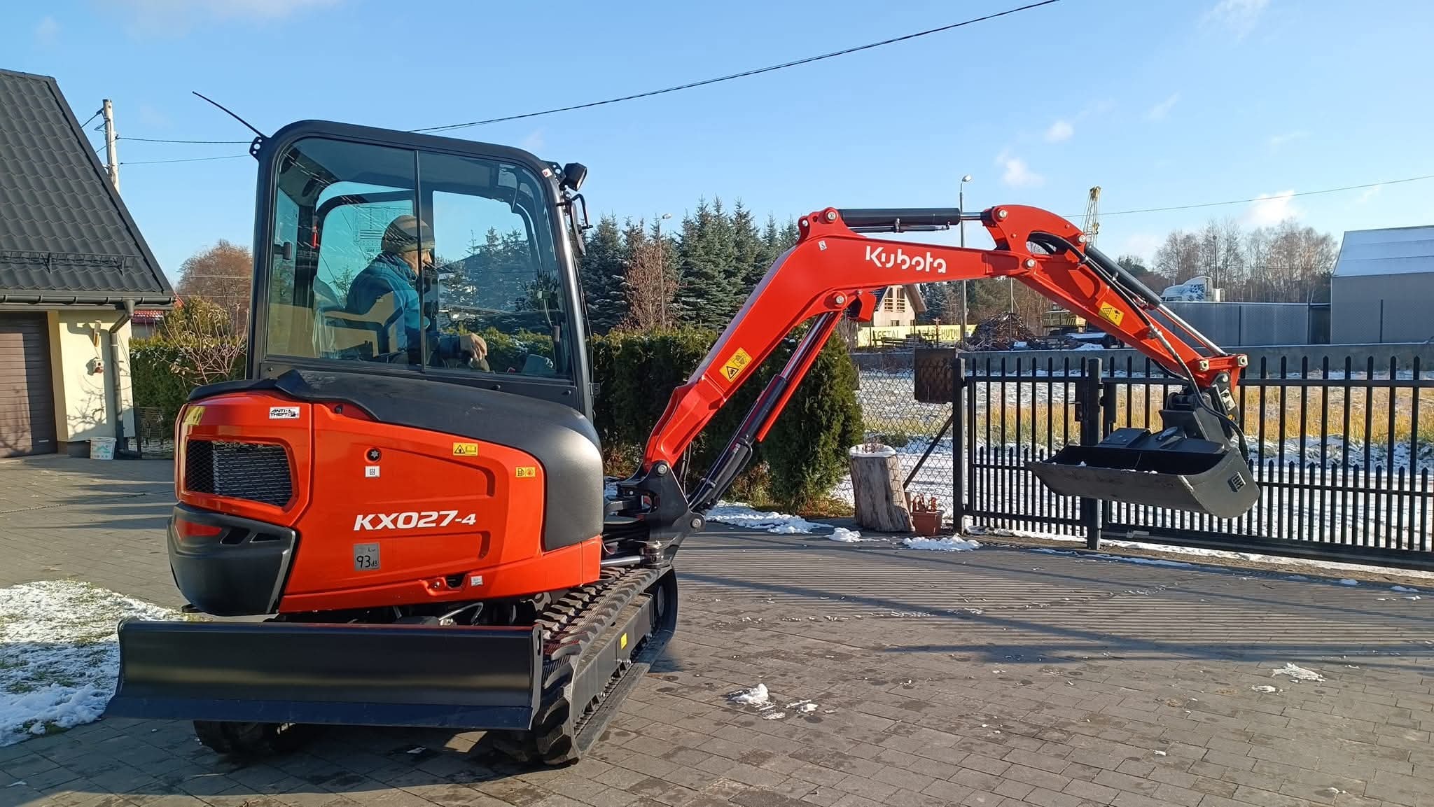 Czerwona minikoparka Kubota KX027-4 z operatorem w kabinie, stojąca na kostce brukowej przed czarnym ogrodzeniem, w tle drzewa i budynki w słoneczny dzień.