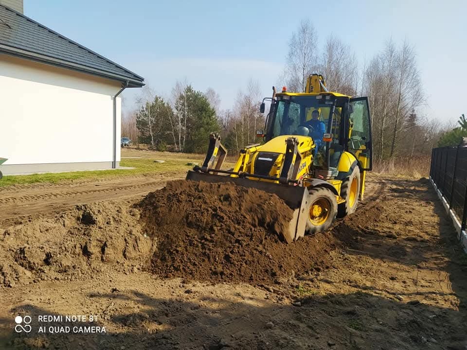 Żółta koparko-ładowarka JCB z otwartymi drzwiami, pracująca na placu budowy przy domu. Widoczna sterta ziemi, ślady opon i operator w kabinie.
