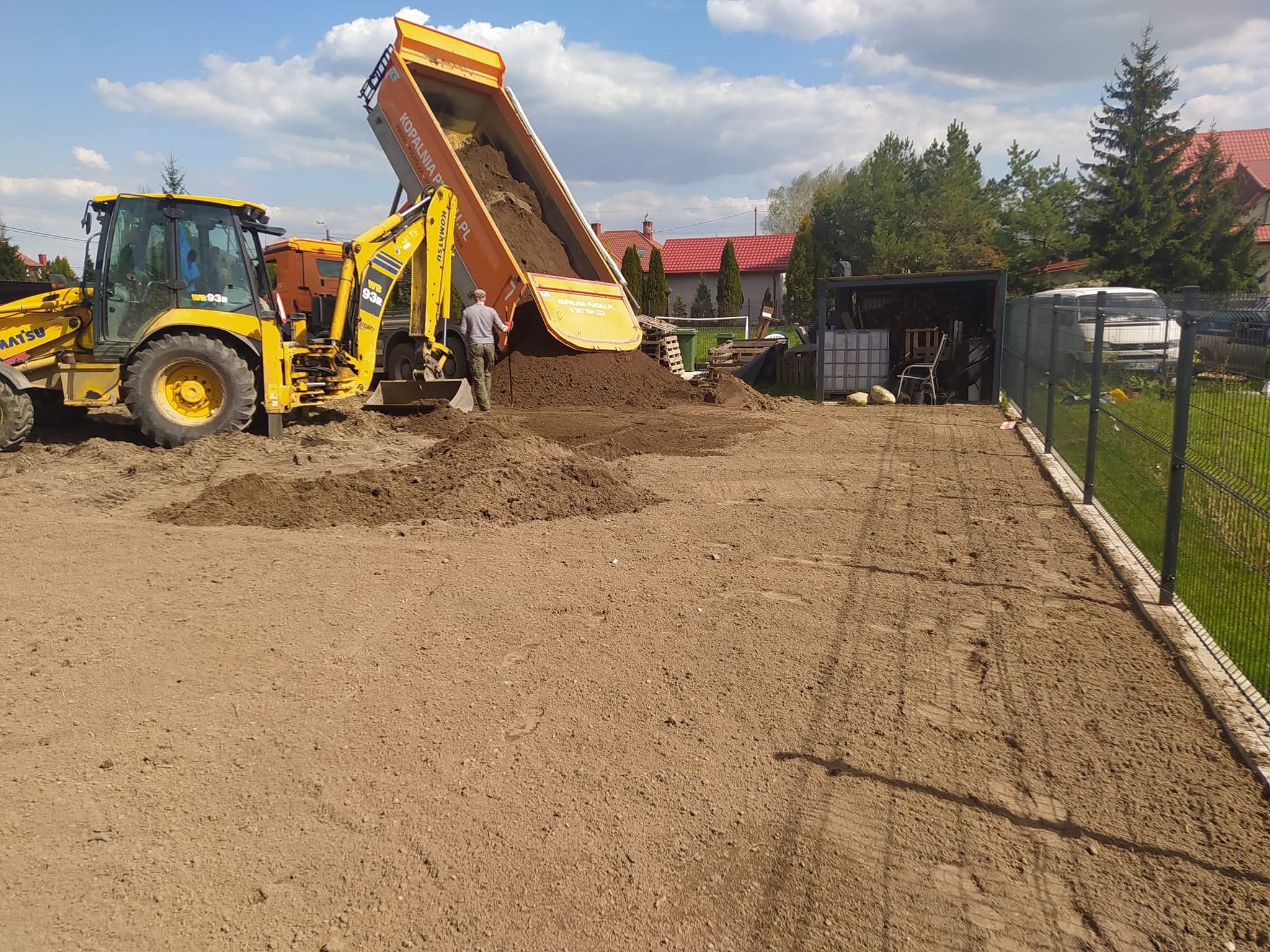 Żółta koparko-ładowarka Komatsu i pomarańczowa wywrotka z uniesioną skrzynią wysypują ziemię na wyrównany teren, widoczny pracownik w tle, ogrodzenie z siatki.