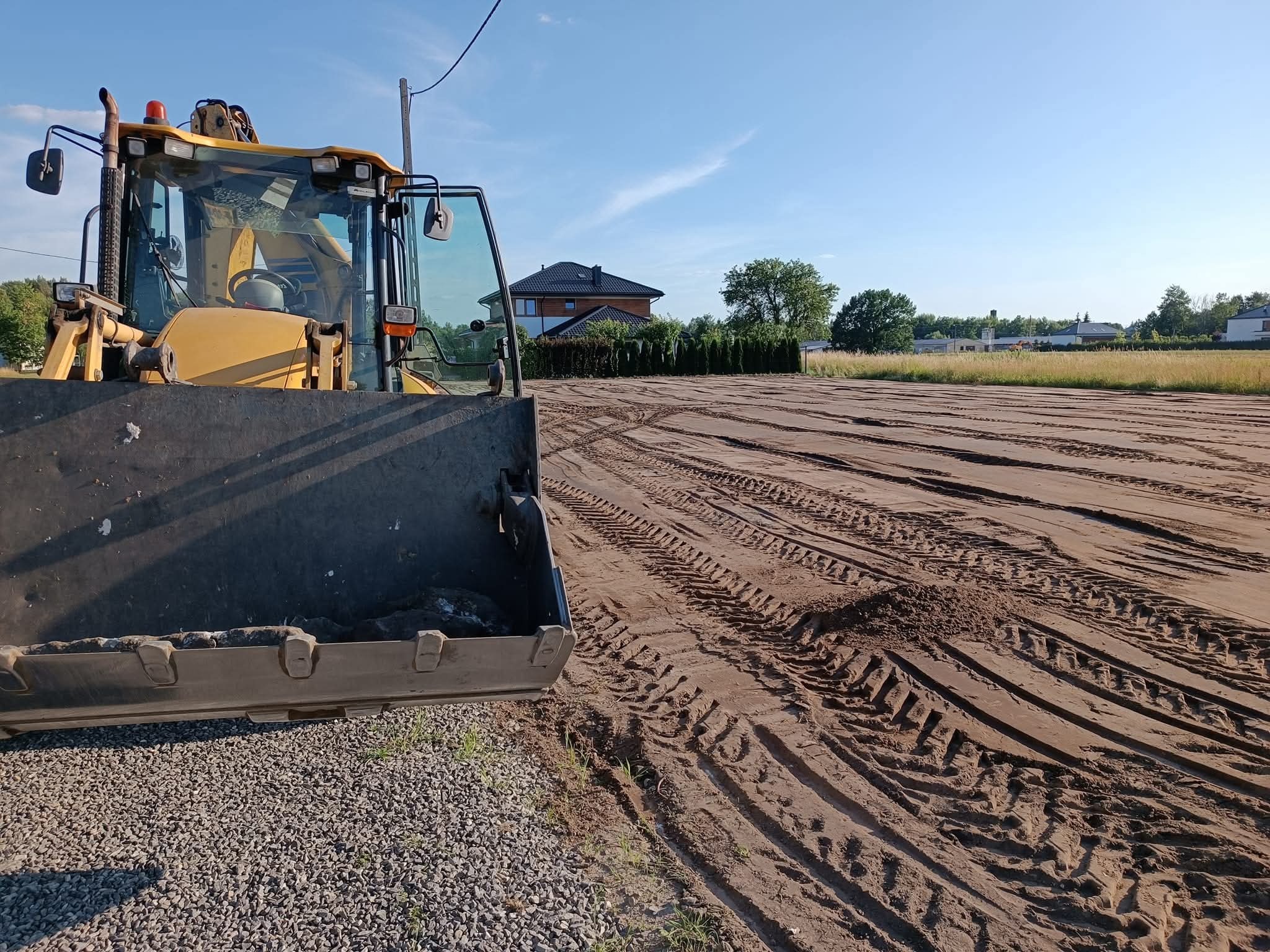 Żółta koparko-ładowarka na działce z wyrównanym, brązowym gruntem. Widoczne ślady opon, w tle domy i drzewa pod błękitnym niebem.