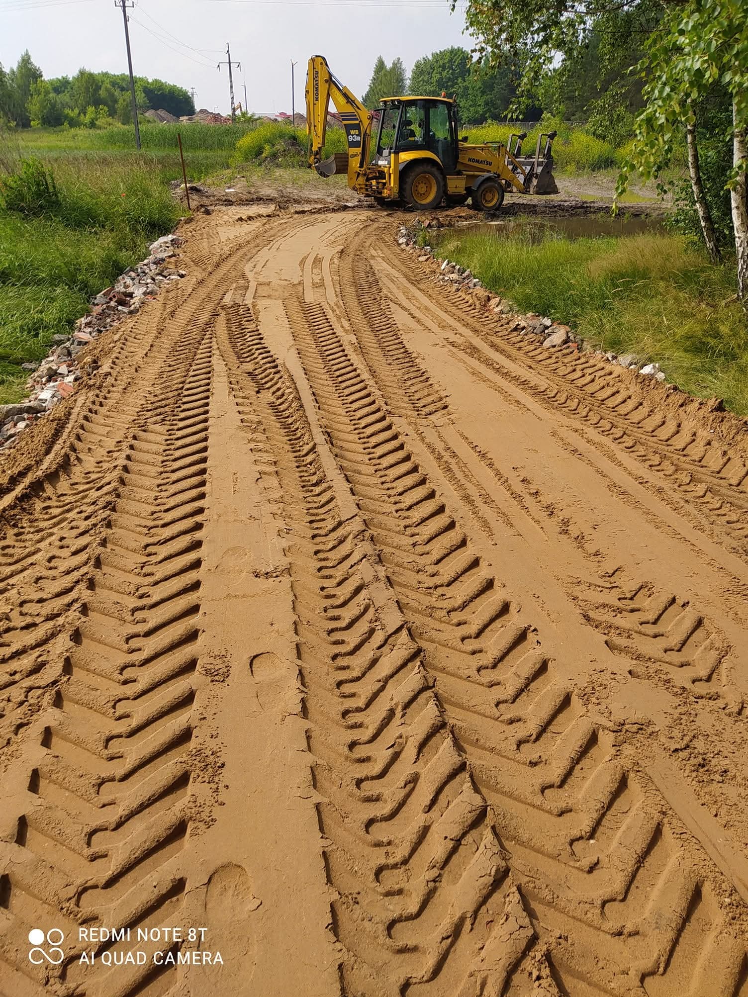 Żółta koparko-ładowarka Komatsu na piaszczystej drodze z wyraźnymi śladami opon. Po bokach gruz i roślinność. Słoneczny dzień, widok z perspektywy.
