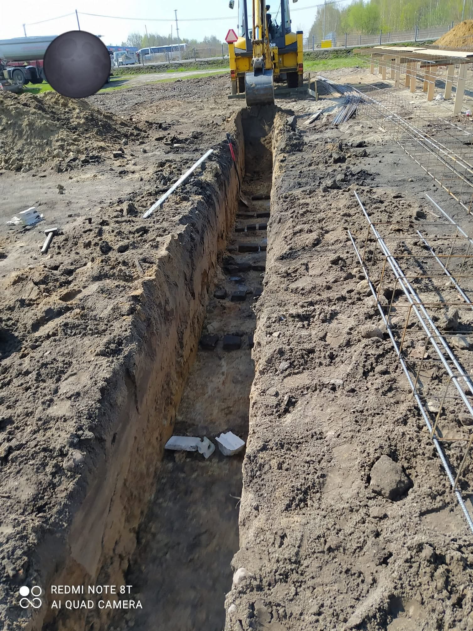 Głęboki wykop w ziemi, z widocznym żółtym koparko-ładowarką i elementami zbrojenia. Widok z góry na przygotowany rów pod instalacje lub fundamenty.