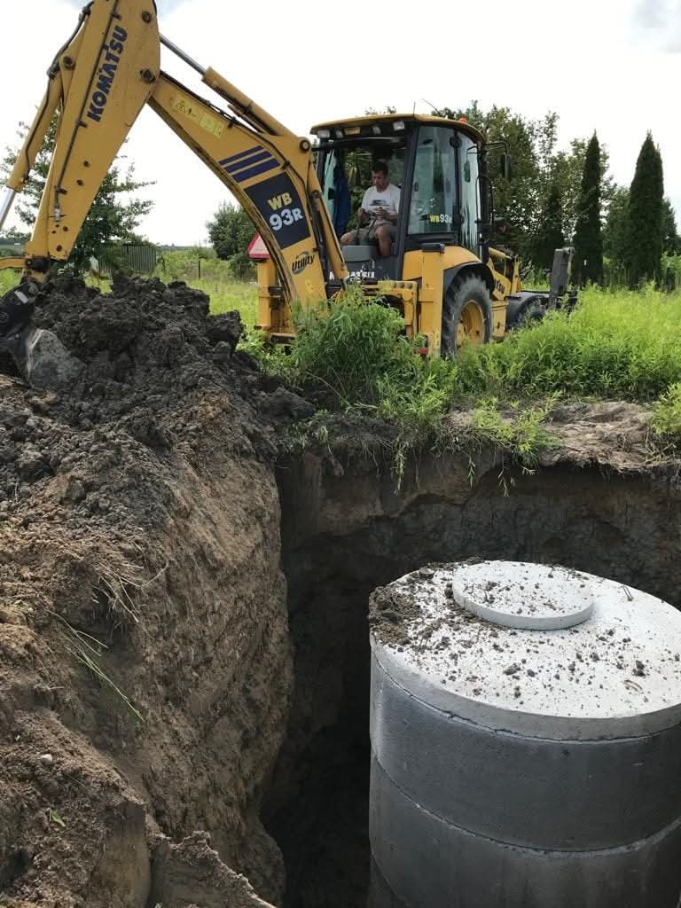 Żółta koparko-ładowarka Komatsu przy wykopie z betonowym szambem; widoczny operator w kabinie, świeżo usypana ziemia i zieleń w tle, pochmurny dzień.