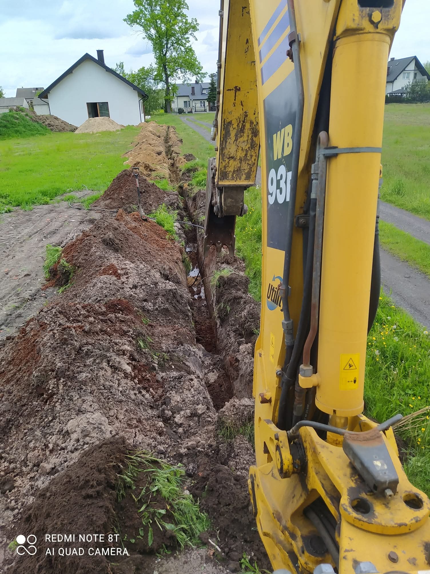 Żółta koparka WB93R w trakcie wykopu wąskiego rowu w ziemi na tle zieleni i zabudowań. Widoczny fragment wykopu z ułożonymi kablami.
