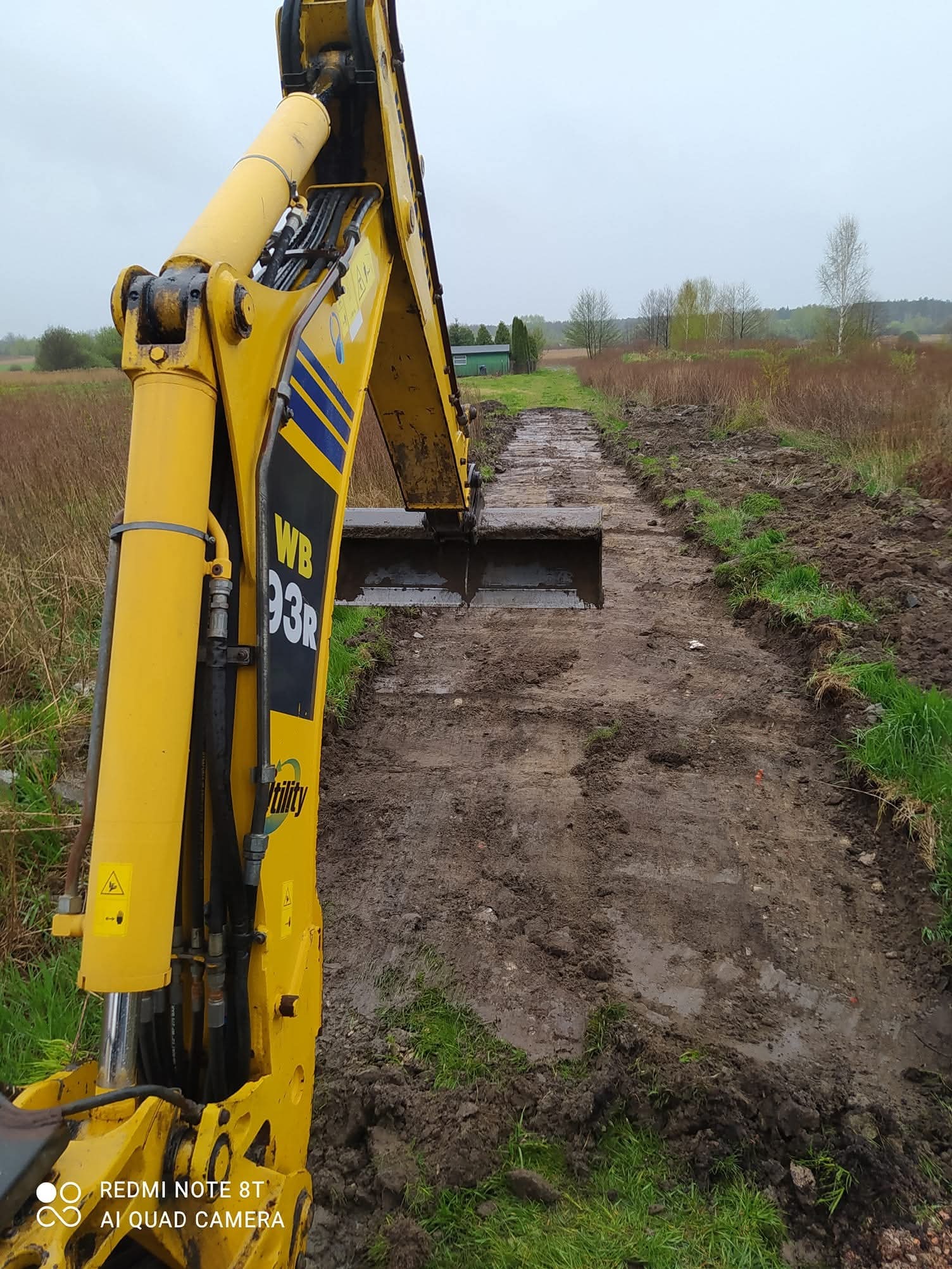 Żółta koparka WB 93R częściowo widoczna, kopiąca wąski pas ziemi na zewnątrz, w tle łąka i drzewa. Ślady błota na ziemi.