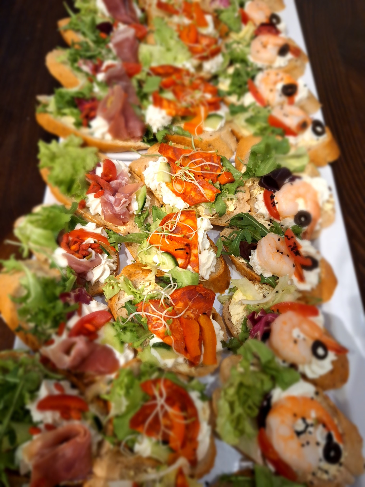 Apetyczne, różnorodne kanapki na białej tacy: krewetki, szynka parmeńska, warzywa. Idealne na catering.