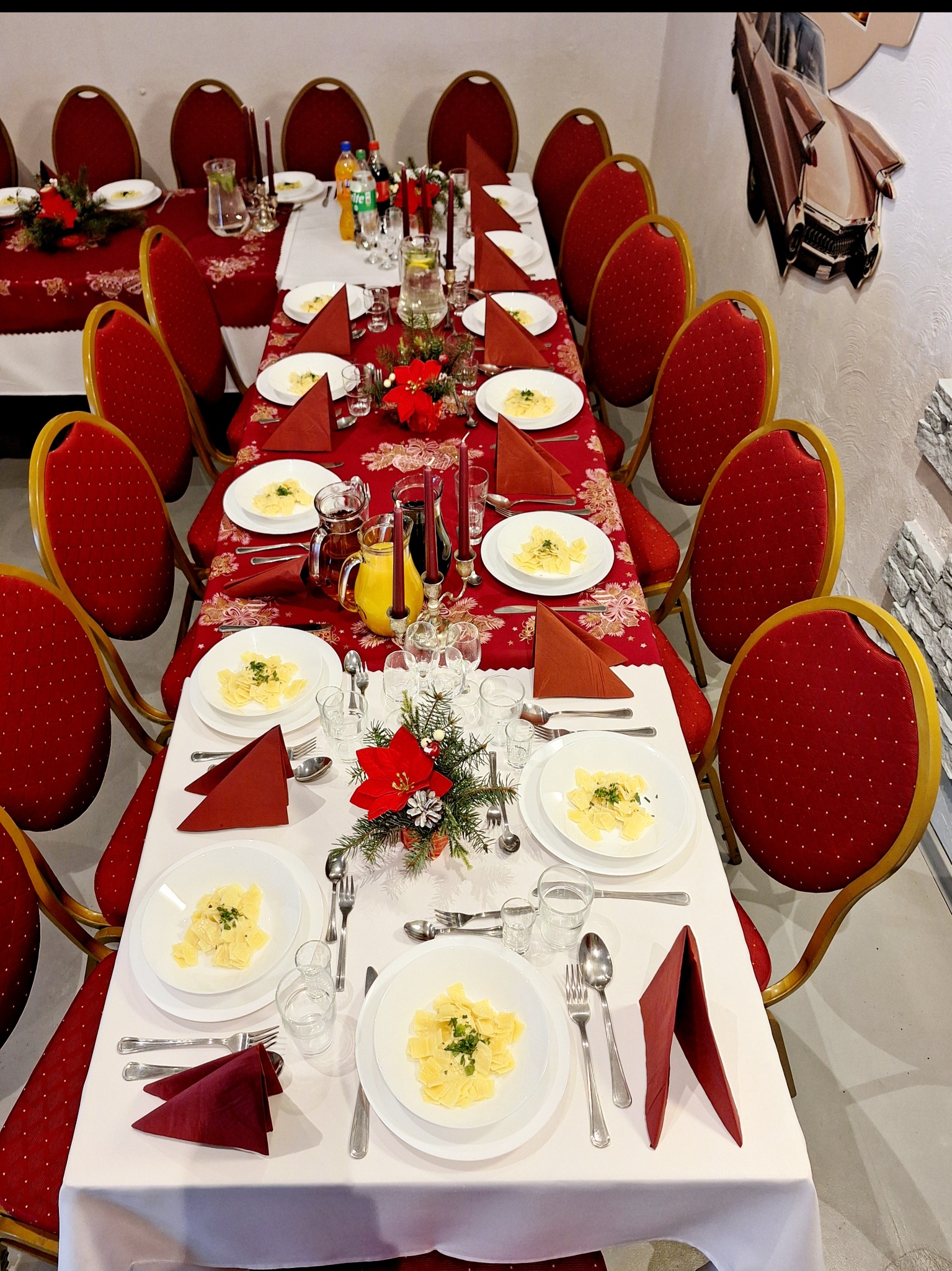 Uroczysty stół wigilijny z czerwonym obrusem w kwiaty, białymi talerzami z daniami i dekoracjami z poinsecji, nakryty dla wielu gości, z krzesłami z czerwonym obiciem.