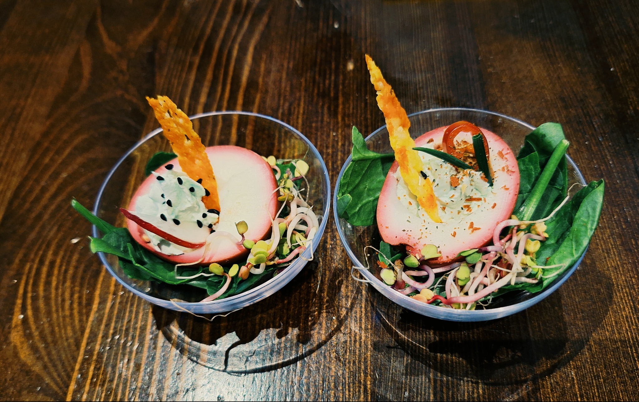 Dwie porcje wykwintnej przekąski w przezroczystych miseczkach na drewnianym stole. Sałata, kiełki, plasterek rzodkiewki i chrupiący dodatek.