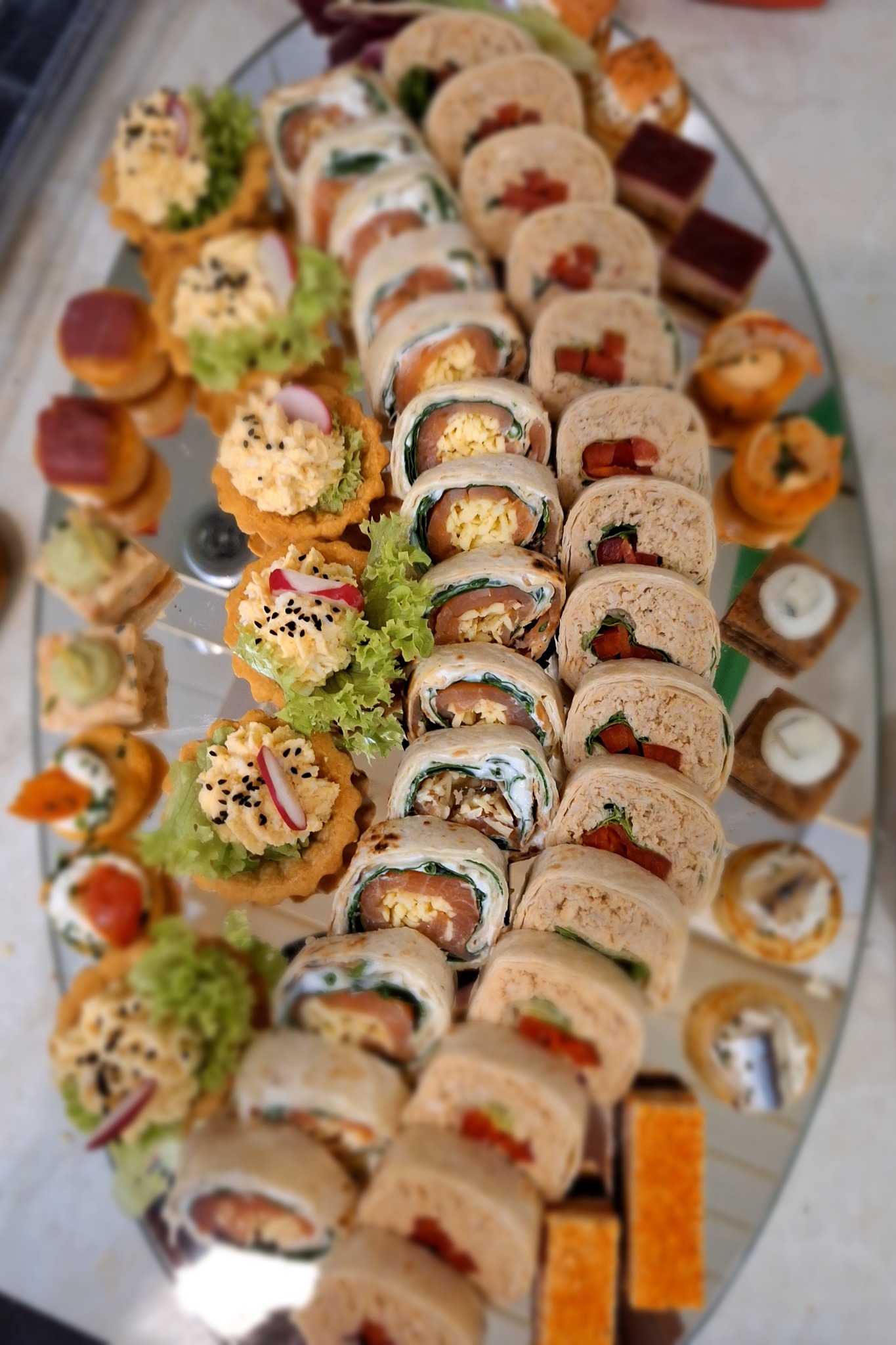 Elegancki wybór przekąsek cateringowych na szklanej tacy: tartaletki z sałatką, roladki z tortilli i mini kanapeczki. Różnorodność smaków i estetyczne podanie.