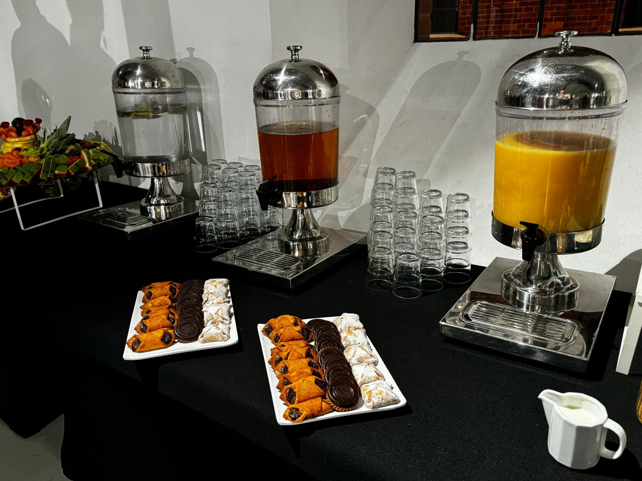 Elegancki bufet z napojami (woda, herbata, sok pomarańczowy) w dyspenserach i słodkimi przekąskami na czarnym obrusie, przygotowany na przyjęcie.