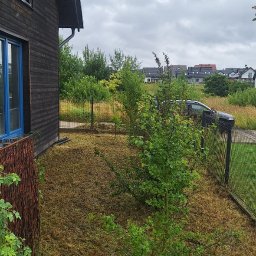 Damian Popławski - Ogród po koszeniu trawy, widoczne ścięte źdźbła. Drewniany dom z niebieskimi oknami w tle. Metalowe ogrodzenie oddziela posesję od drogi. Pochmurne niebo.