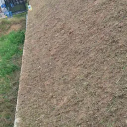 Świeżo skoszona trawa na zboczu z siatką ogrodzeniową w tle. Widoczne domy i drzewa. Częściowo widoczny trawnik z dłuższą trawą.