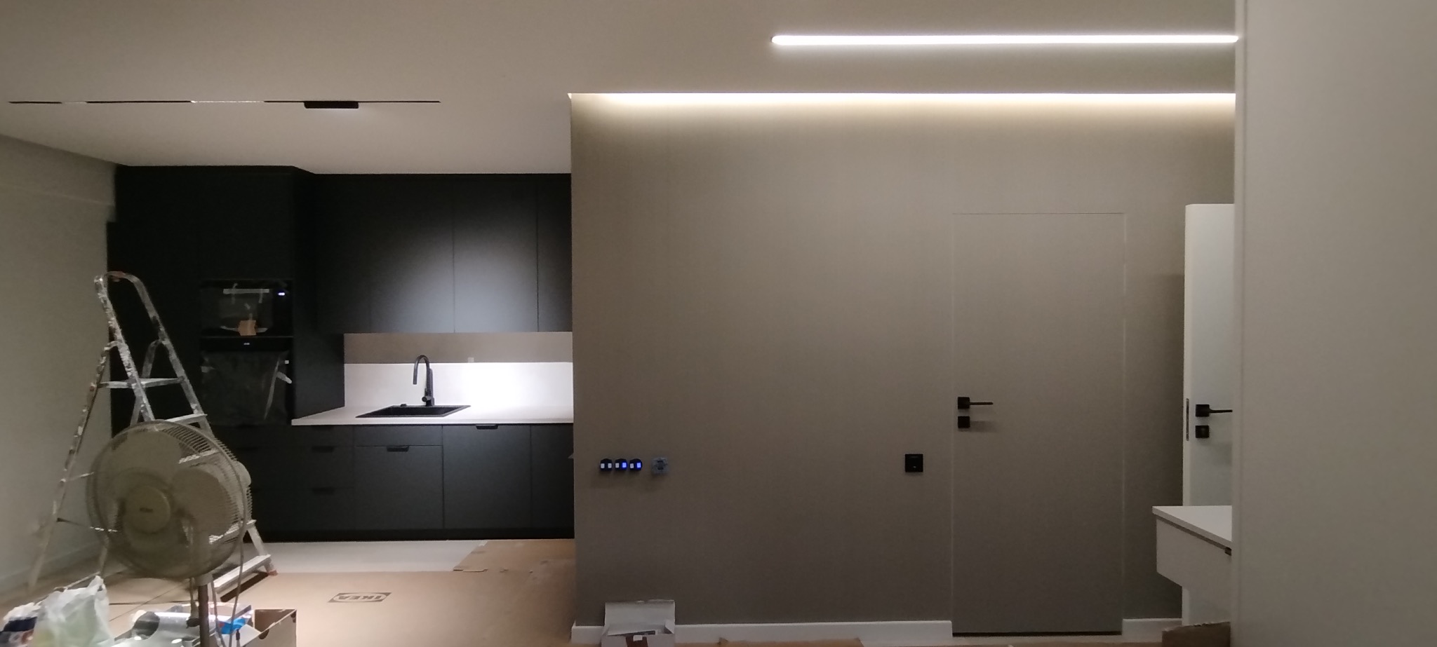 Nowoczesna kuchnia z czarną zabudową i białym blatem, w trakcie wykończenia. Widoczna drabina i wentylator. Minimalistyczny design, oświetlenie LED.